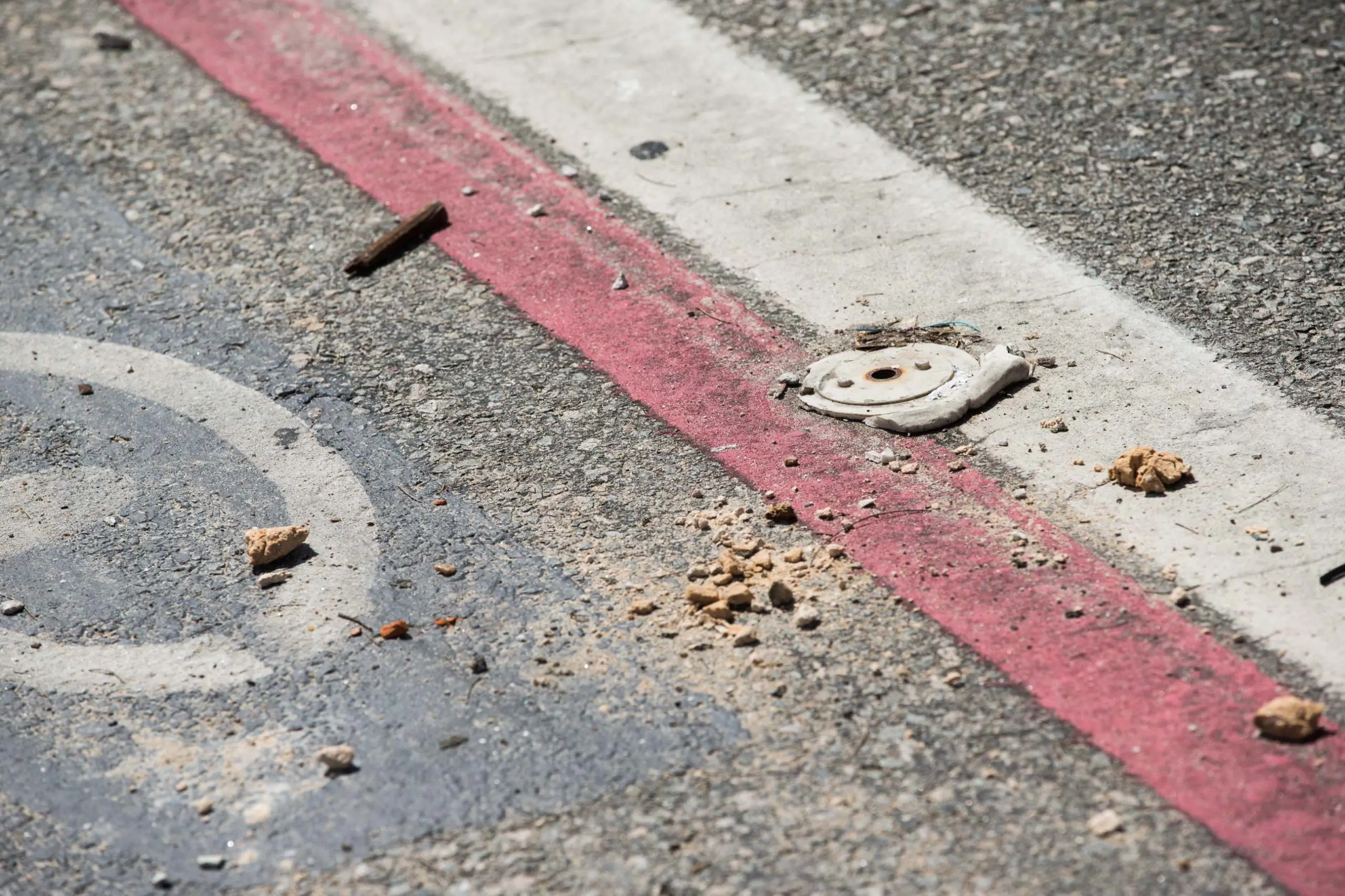 Um close-up do asfalto desgastado, focando em uma linha vermelha de sinalização e na base metálica de um balizador que foi arrancado, restando apenas detritos e poeira ao redor.