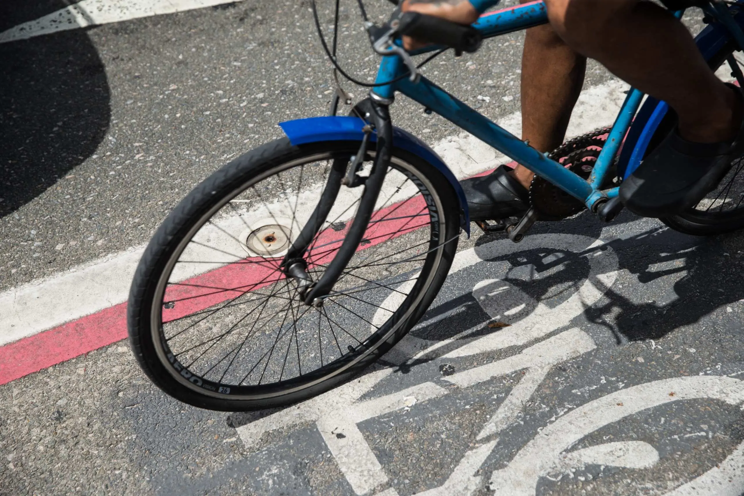 Um ciclista trafega sobre uma ciclofaixa asfáltica que possui sinalização branca de uma bicicleta e uma linha delimitadora vermelha e branca. A imagem foca no pneu dianteiro preto, no quadro azul da bicicleta e nos pés do ciclista, que calça sapatos pretos fechados. A cena é iluminada pelo sol, projetando sombras nítidas no chão.