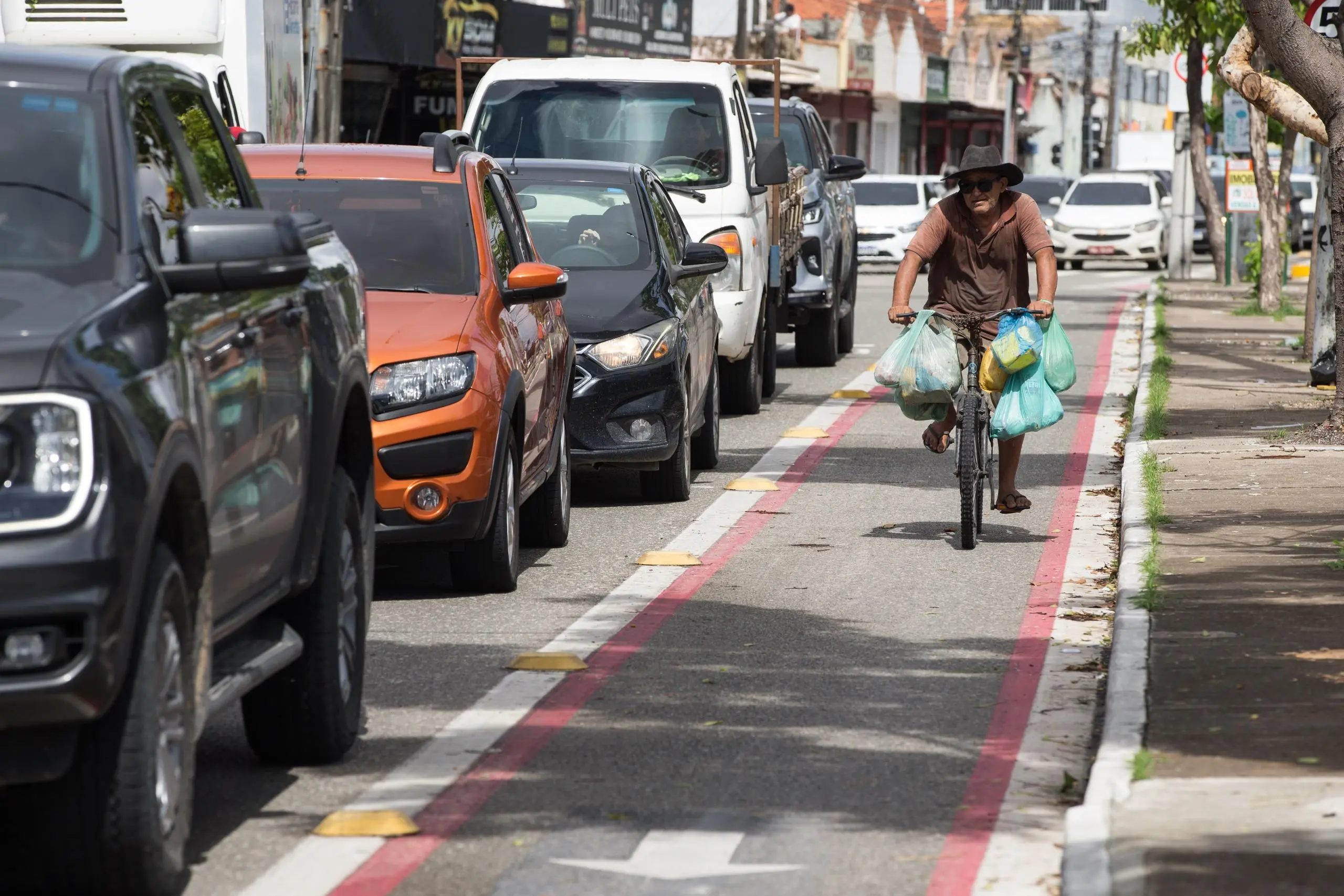 Um ciclista de chapéu e óculos escuros pedala por uma ciclofaixa delimitada por tachões amarelos e linhas vermelhas, carregando diversas sacolas plásticas penduradas no guidão. À esquerda, uma fila de carros parados em um congestionamento urbano acompanha o trajeto, enquanto à direita vê-se uma calçada com árvores. A cena ocorre durante o dia sob luz solar intensa, destacando o contraste entre a agilidade da bicicleta e o trânsito estagnado.