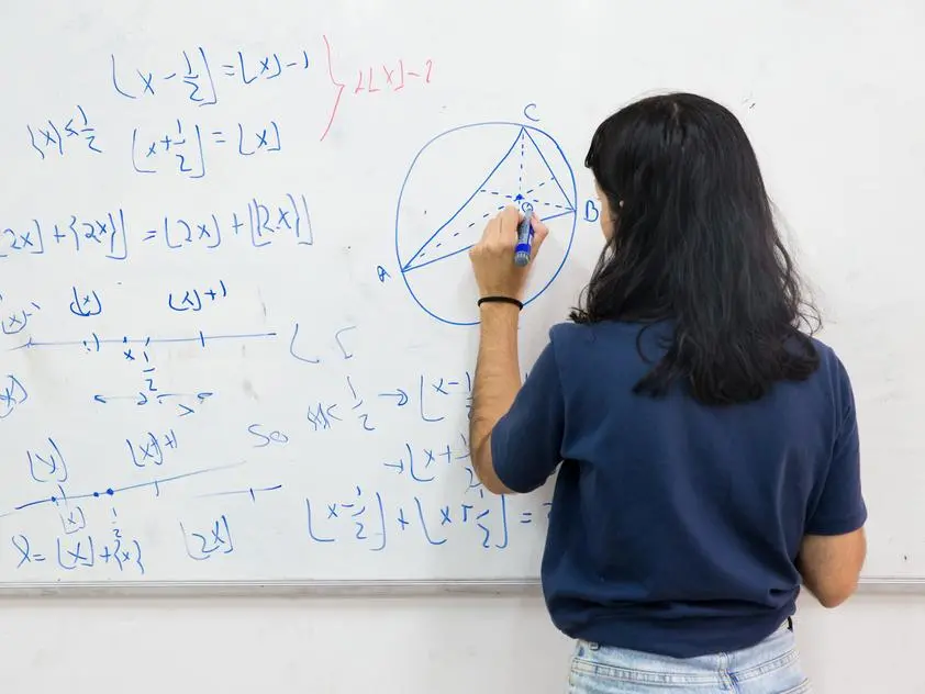 Professora vista de costas escreve em um quadro branco com fórmulas matemáticas e um desenho geométrico de um triângulo inscrito em um círculo, com pontos identificados por letras.