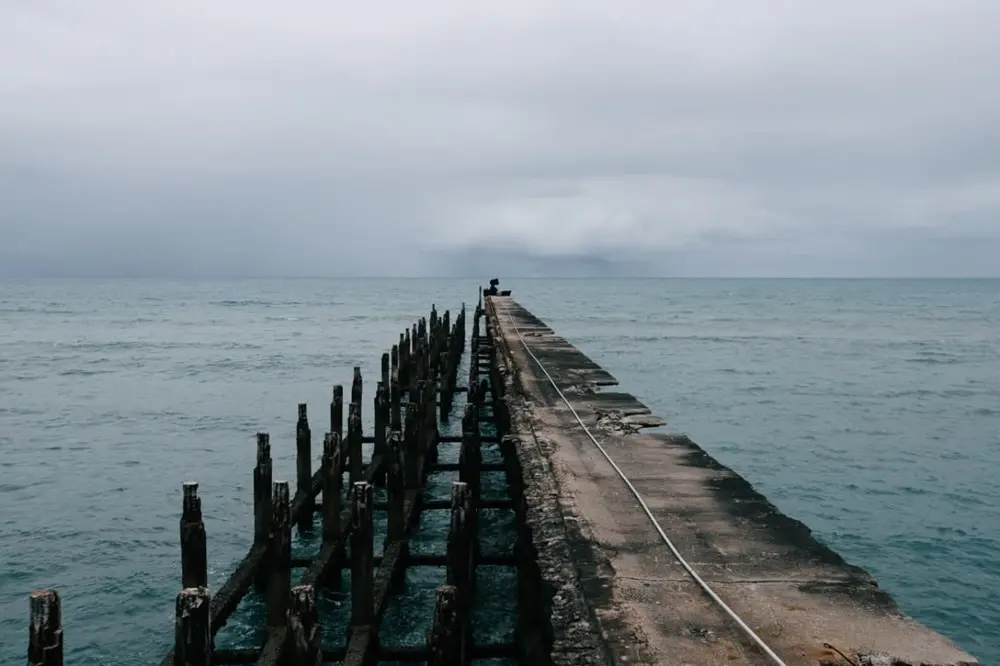 Na imagem, fotografia em plano geral e perspectiva profunda. Um antigo píer de concreto, estreito e desgastado, estende-se em linha reta até o horizonte. No final dele, a pequena silhueta da escultura 