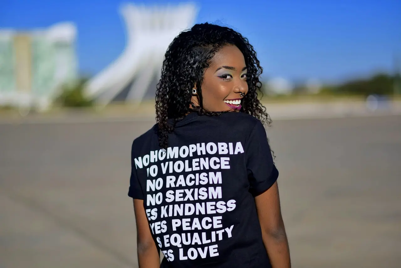 Mulher negra com cabelos cacheados olha para trás e sorri enquanto veste uma camiseta preta com frases contra homofobia, racismo, sexismo e violência e a favor de paz, igualdade e amor. Ao fundo, desfocado, aparece a Catedral de Brasília.