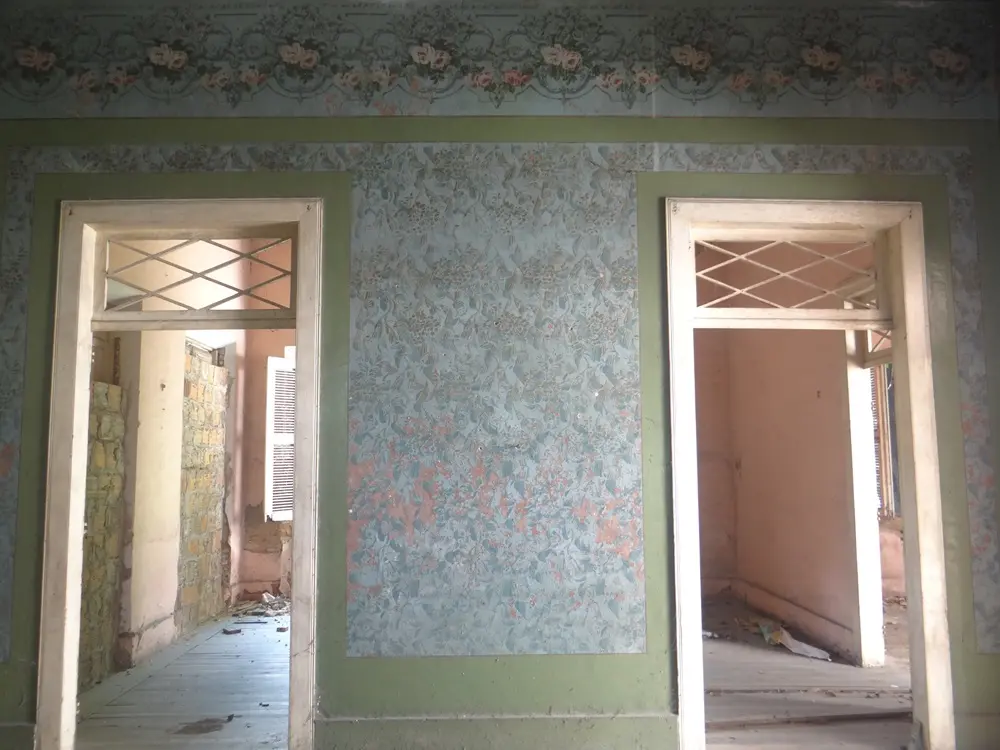 Na imagem, vista interna de uma casa histórica em estado de abandono, mostrando duas aberturas de portas lado a lado. A parede entre as portas possui um papel de parede azul com padrões florais desgastados, emoldurado por uma pintura verde. Acima, há uma faixa decorativa (barrado) com flores rosadas. Através das portas, veem-se cômodos vazios com assoalho de madeira, paredes descascadas, tijolos expostos e luz natural vindo de janelas ao fundo. O teto das passagens possui detalhes em madeira em formato de X.