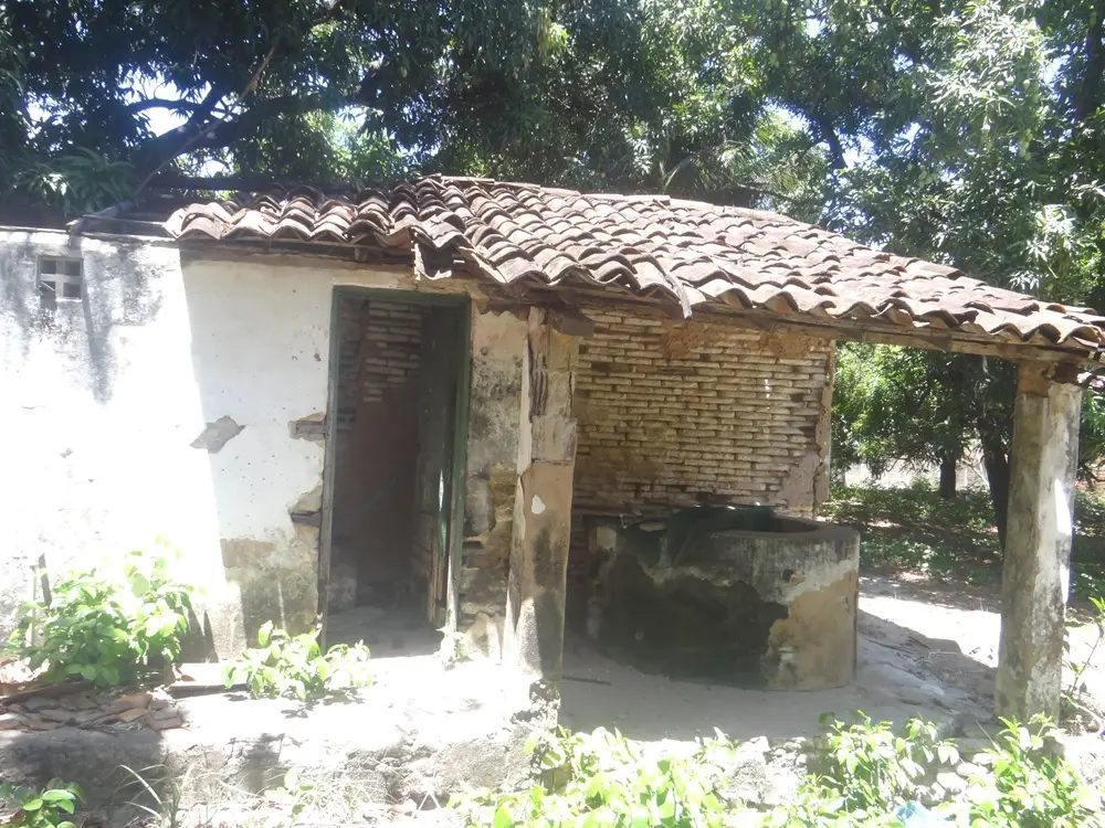 Na imagem, fotografia de uma pequena construção rústica e anexa, em avançado estado de deterioração, localizada em uma área arborizada. A estrutura possui um telhado de telhas de barro canal, parcialmente desmoronado, sustentado por pilares simples. À esquerda, uma parede branca descascada com uma porta de madeira verde aberta; à direita, uma parede de tijolos aparentes sem reboco. No centro, sob a cobertura, há uma estrutura circular de cimento que se assemelha a um poço ou tanque antigo. O entorno é composto por terra batida e árvores frondosas que projetam sombras sobre a construção.
