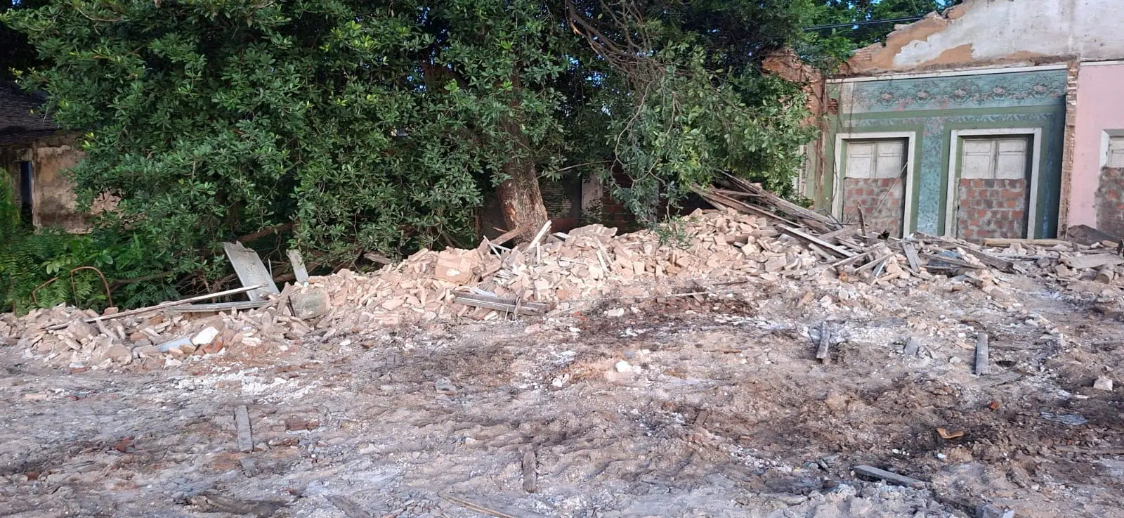 Na imagem, fotografia de uma área de demolição onde se veem os escombros de uma casa histórica. No primeiro plano, o chão é coberto por terra batida, pedaços de madeira e restos de alvenaria. Ao fundo, à direita, resta apenas uma parede externa em pé, exibindo o mesmo papel de parede azul floral e molduras verdes vistos em fotos anteriores, mas agora com as aberturas de portas ou janelas vedadas com tijolos. À esquerda, uma grande árvore com folhagem densa cobre parte das ruínas. O cenário é de abandono e destruição total da estrutura do imóvel.