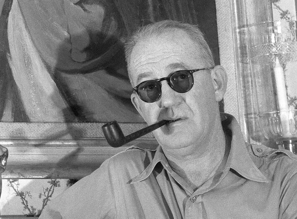 Homem com óculos escuros, barba por fazer, segurando um cachimbo na boca, em ambiente descontraído, com pintura de fundo ao lado de uma janela.