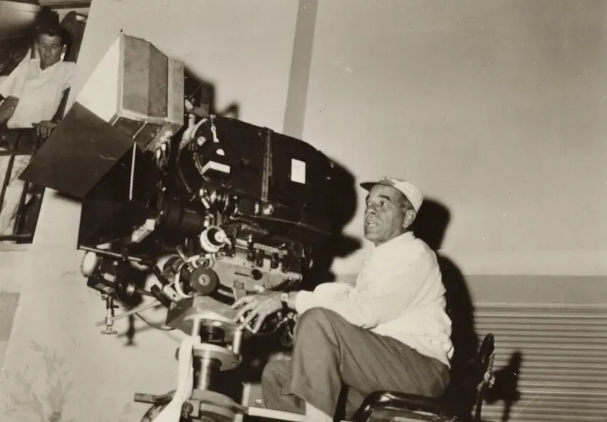 Fotografia antiga de um homem operando uma câmera de cinema em um estúdio, com outros equipamentos ao fundo. Imagem em preto e branco, mostrando a história do cinema brasileiro.