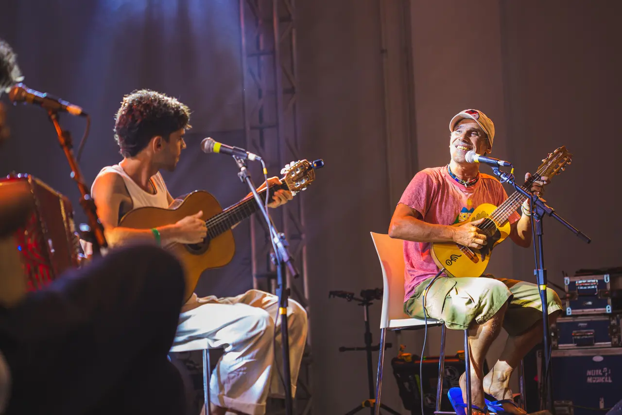 Manu Chao sorri e toca um pequeno violão amarelo enquanto está sentado no palco, acompanhado pelo filho Kirá, também ao violão, em uma atmosfera descontraída. A iluminação cênica destaca os artistas em uma composição vibrante que captura a energia e a cumplicidade da apresentação musical.