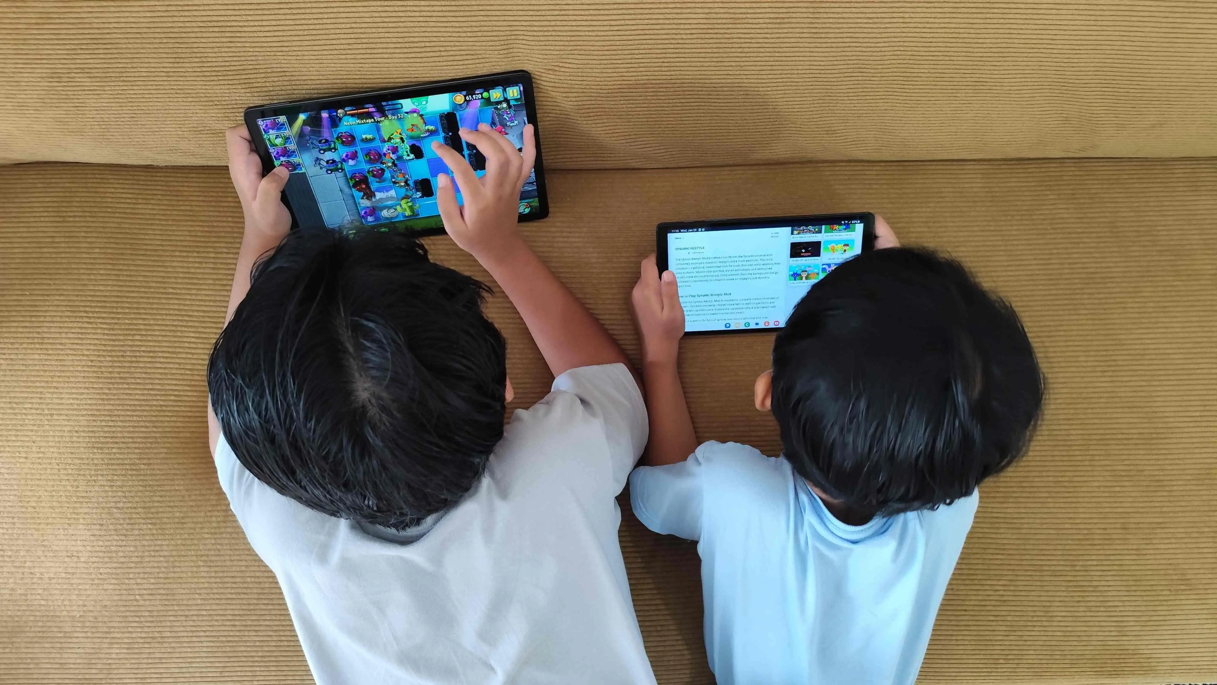 Vista aérea de duas crianças deitadas em um sofá, cada uma usando um tablet. Uma joga um game colorido e a outra navega em uma página de texto. Cena ilustra o consumo digital infantil.