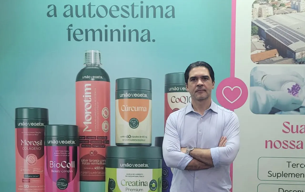 Ronaldo Albuquerque, homem de braços cruzados em frente a um painel publicitário com produtos de suplementos alimentares da marca União Vegetal. No fundo aparecem embalagens de itens como Morotim, Cúrcuma, CoQ10, Morosil, BioColl e Creatina, além de um texto sobre autoestima feminina.