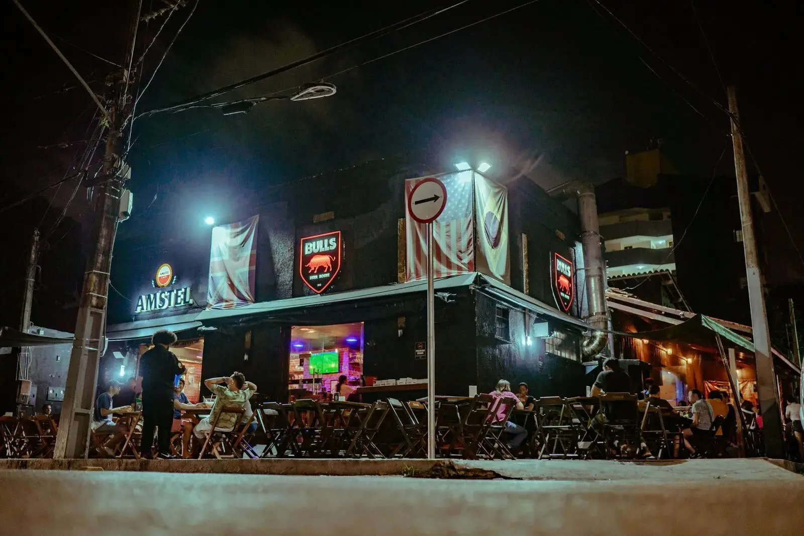 Bares e casas de shows à noite com decoração temática de futebol, incluindo banners do Flamengo e do Botafogo, com pessoas assistindo a uma partida pela TV ao ar livre.