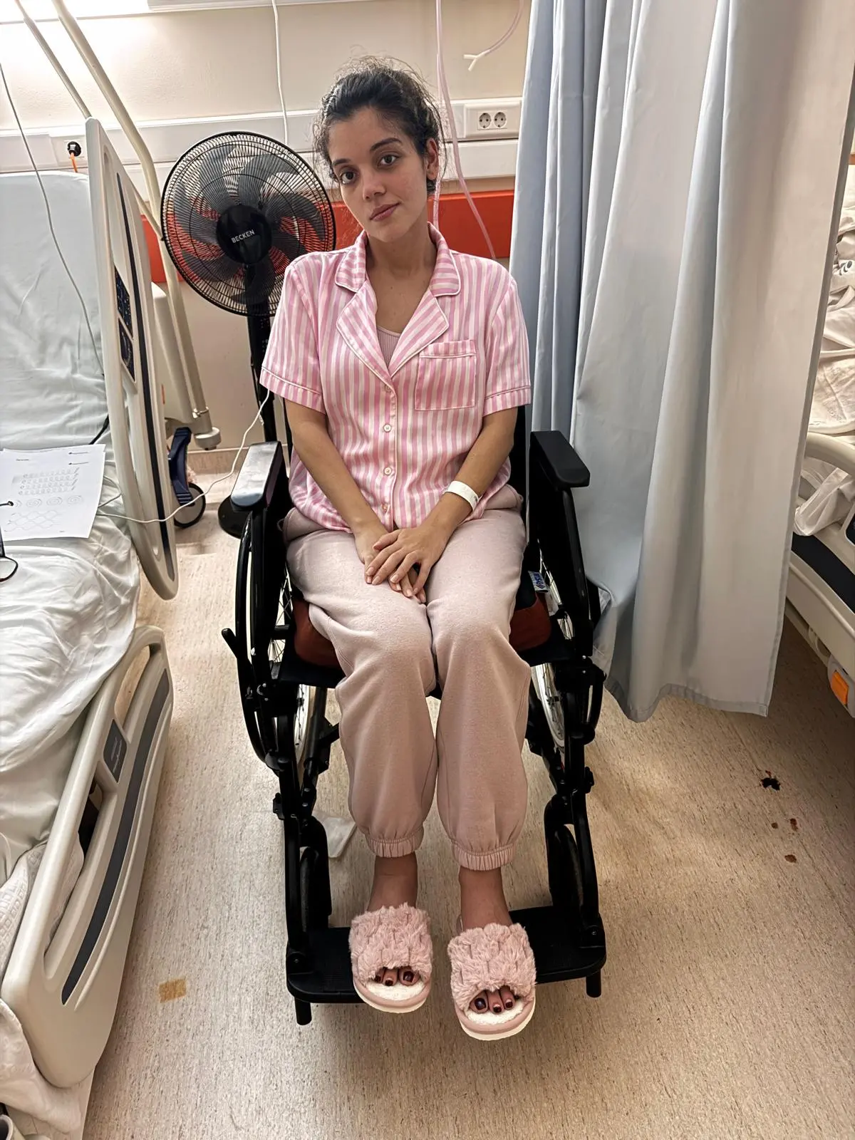 Na imagem, uma mulher jovem de pele clara e cabelos escuros presos sentada em uma cadeira de rodas no que parece ser um quarto de hospital. Ela veste um pijama listrado de rosa e branco, calça de moletom rosa clara e chinelos de pelúcia rosa. Ela olha diretamente para a câmera com uma expressão serena. Ao fundo, veem-se camas hospitalares, um ventilador de coluna preto e cortinas divisórias azuis claras.