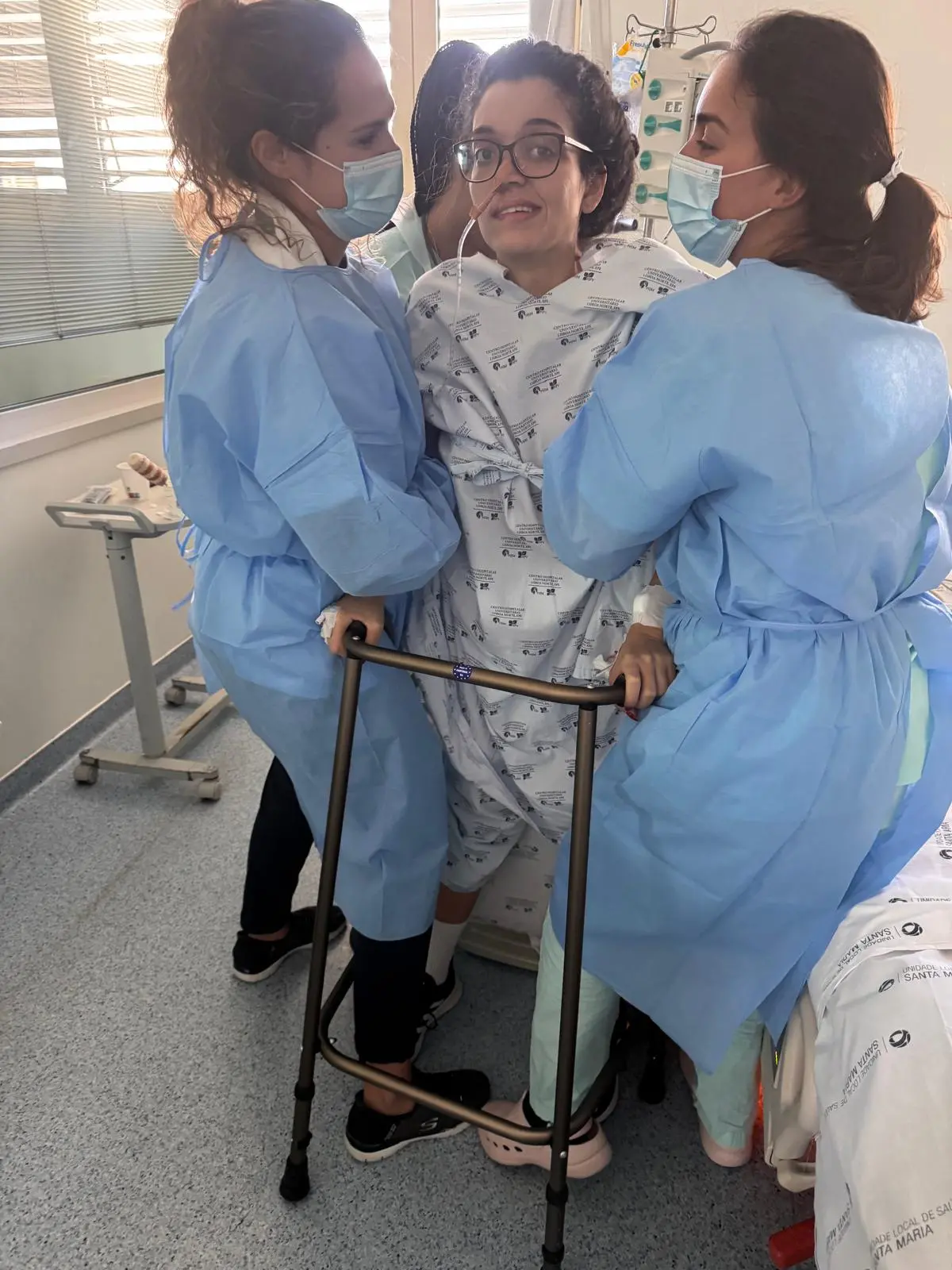 Na imagem, uma mulher jovem, de óculos e cabelos presos, é amparada por duas profissionais de saúde enquanto se levanta pela primeira vez em um quarto de hospital. Ela veste um roupão hospitalar branco com estampas azuis e utiliza um andador de metal para se apoiar. As profissionais estão de costas para a câmera, usando aventais descartáveis azuis e máscaras protetoras. A paciente olha para a frente com um leve sorriso de esforço e determinação. Ao fundo, vê-se a janela do hospital com persianas e equipamentos médicos.