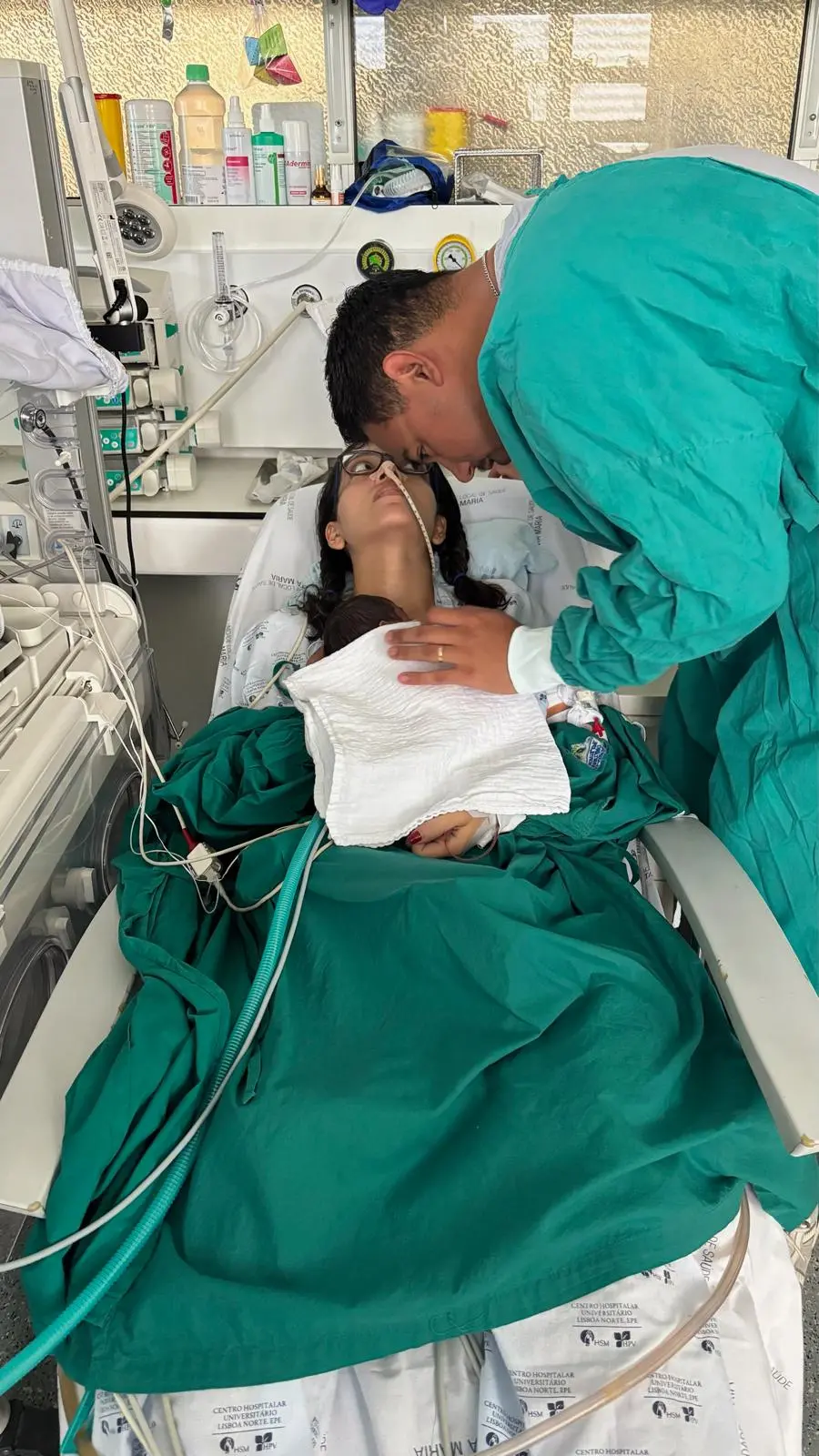 Na imagem, uma mulher jovem está deitada em uma cama de hospital, coberta por um lençol verde, vivendo o primeiro contato com sua filha recém-nascida. O bebê está envolto em uma manta branca sobre o peito da mãe. A mulher usa óculos, tem cabelos escuros presos em tranças e possui um tubo nasal. Um homem vestindo avental cirúrgico verde se inclina sobre elas, encostando suavemente sua testa na da mulher, em um gesto de carinho e apoio. O ambiente é uma unidade hospitalar, com diversos equipamentos médicos, monitores e frascos ao fundo.