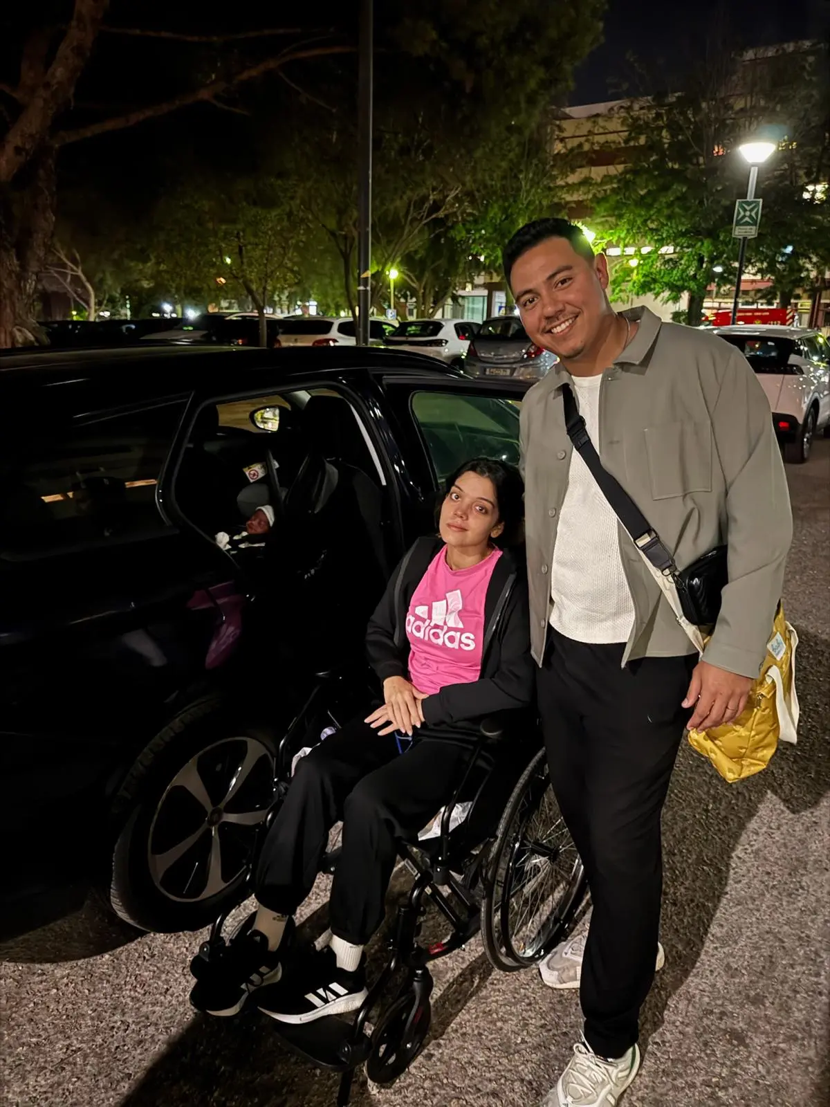 Na imagem, fotografia noturna em um estacionamento. Uma mulher jovem está sentada em uma cadeira de rodas ao lado da porta aberta de um carro preto; ela veste uma camiseta rosa da Adidas, casaco e calças pretas. Dentro do carro, no banco traseiro, vê-se um bebê recém-nascido em uma cadeirinha. Ao lado da mulher, um homem jovem sorri para a câmera; ele veste uma jaqueta cinza sobre uma camiseta branca, calças pretas e carrega uma bolsa amarela a tiracolo. Ao fundo, há árvores, outros carros estacionados e a iluminação pública da rua.