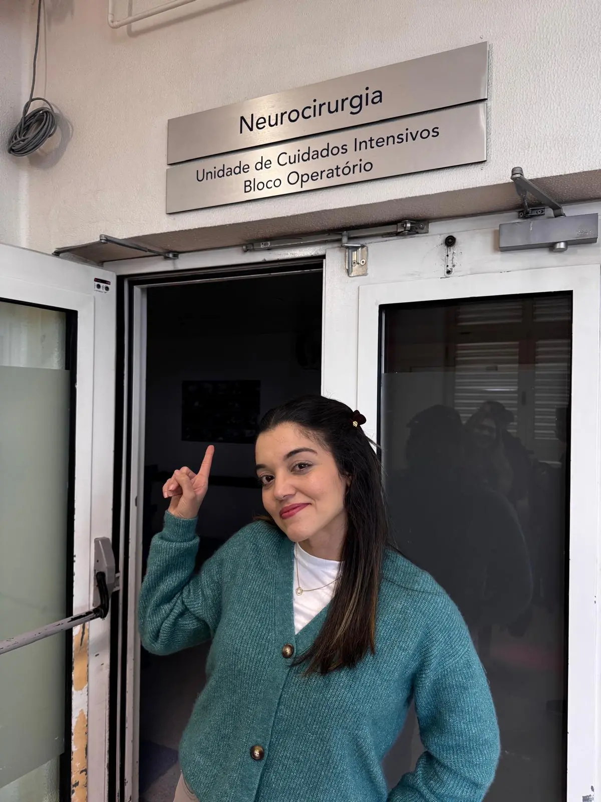 Na imagem, uma mulher jovem de cabelos escuros e leve sorriso posa em frente a uma porta de vidro dupla em um hospital. Ela veste um cardigã de tricô verde sobre uma camiseta branca e aponta com o dedo indicador para cima, em direção a uma placa metálica na parede. A placa contém as inscrições: 