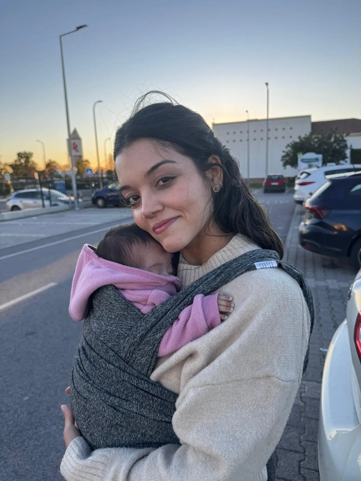 Na imagem, uma mulher jovem de cabelos escuros e expressão serena sorri levemente para a câmera em um ambiente externo ao entardecer. Ela carrega um bebê pequeno junto ao peito em um sling de tecido cinza mesclado. O bebê veste um casaco de moletom rosa com capuz e parece dormir tranquilamente. A mulher usa uma blusa de tricô bege de gola alta. Ao fundo, vê-se um estacionamento com carros, postes de iluminação e o céu com tons suaves de laranja e azul típicos do pôr do sol.