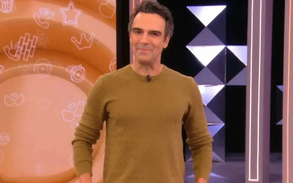 Apresentador de programa de TV sorrindo, vestido com suéter marrom, em estúdio de televisão com fundo decorativo moderno. Ideia para conteúdo de mídia e entretenimento.