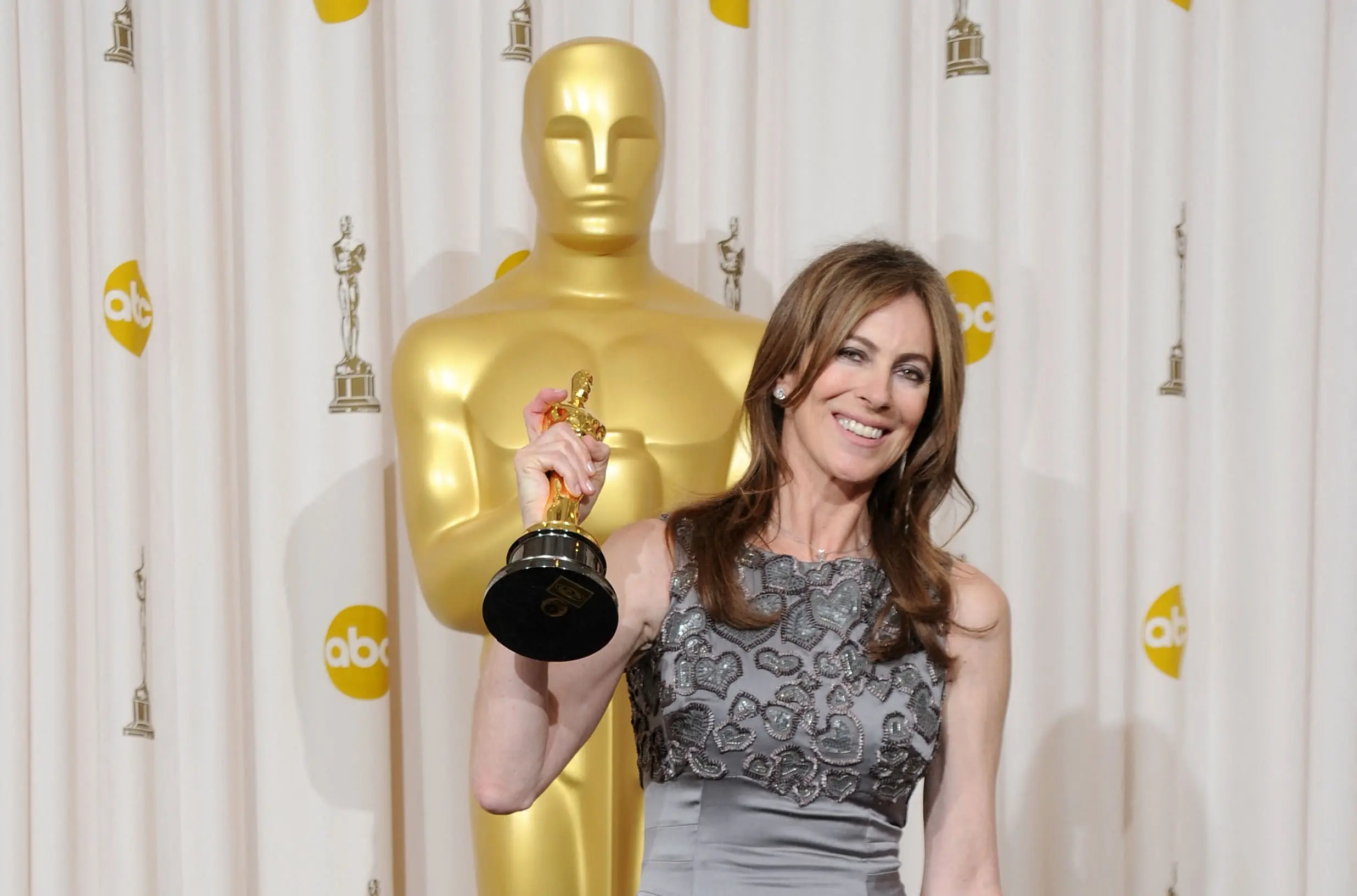 Kathryn Bigelow premiada com um Oscar, sorridente, segurando seu troféu na cerimônia do Oscar 2010, com fundo de cortina branca e estatueta dourada ao fundo.