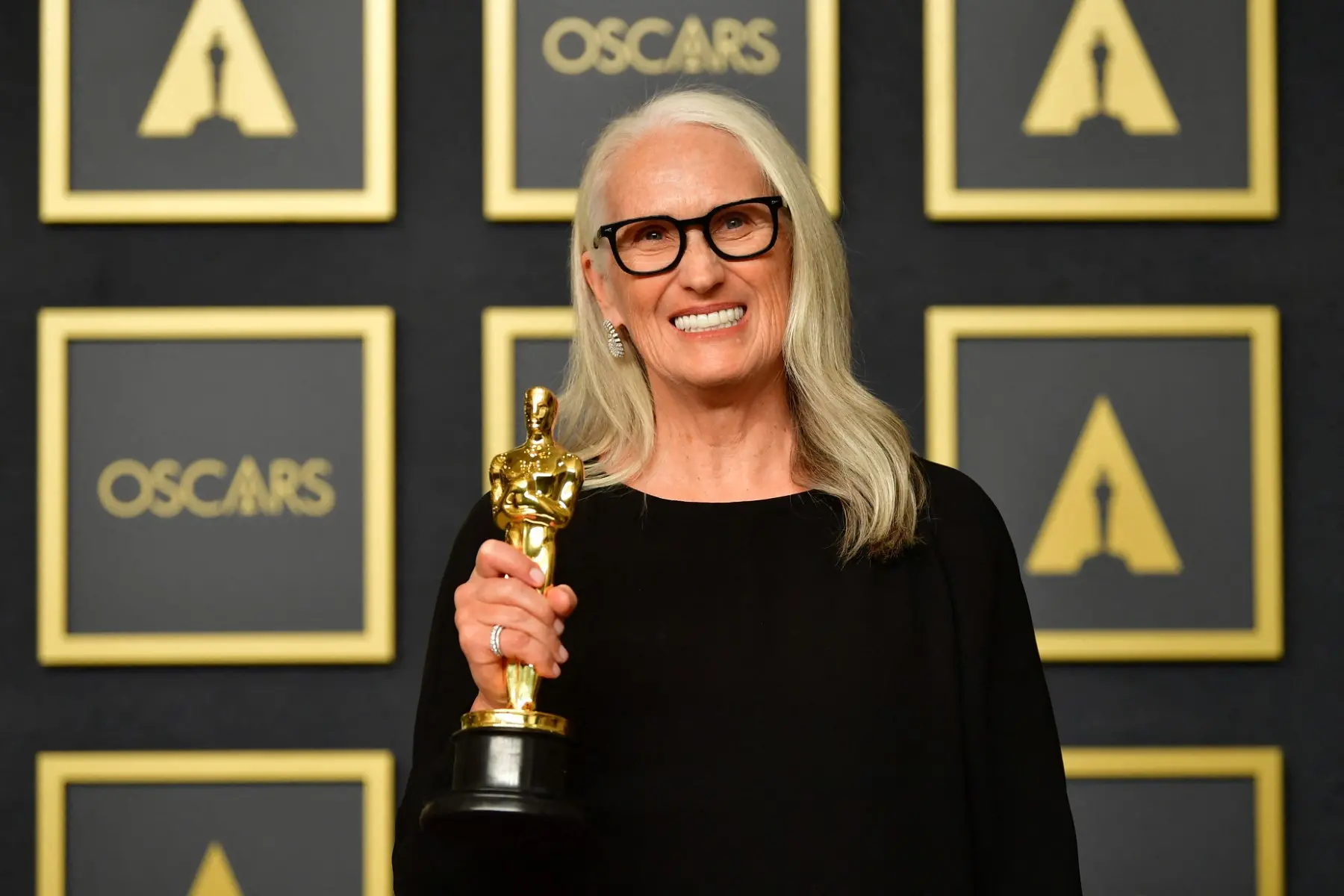Mulher de cabelos longos grisalhos, óculos escuros, sorrindo e segurando um Oscar após ganhar na cerimônia de Oscars, com fundo de painéis com logos de Oscar.