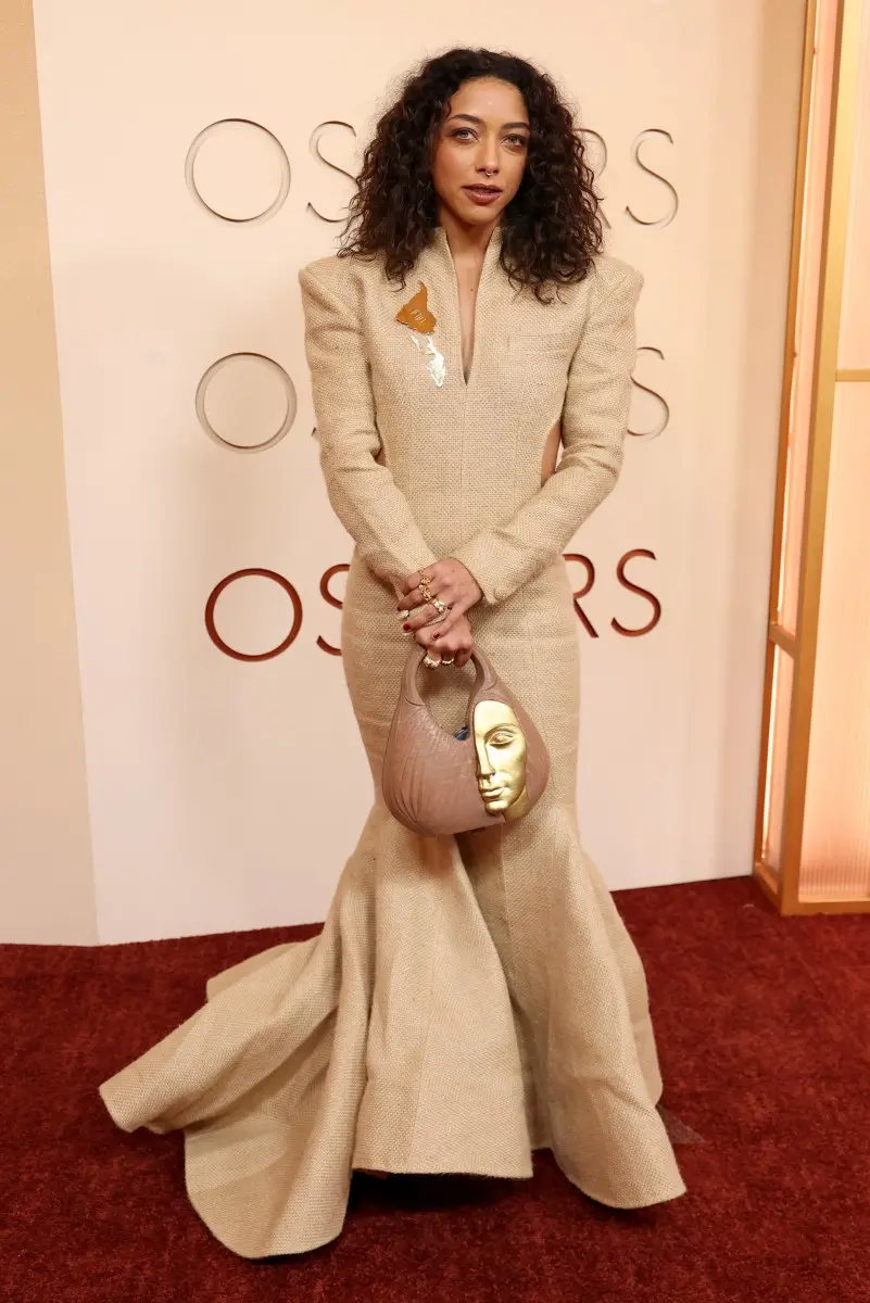 Pessoa em um tapete vermelho posando diante de um painel com a palavra “OSCARS”. A pessoa veste um vestido longo estruturado em tom bege, com ombros marcados, mangas compridas e saia volumosa que se espalha pelo chão. O tecido tem textura semelhante a brocado. No lado do peito há um detalhe metálico decorativo em forma de borboleta.