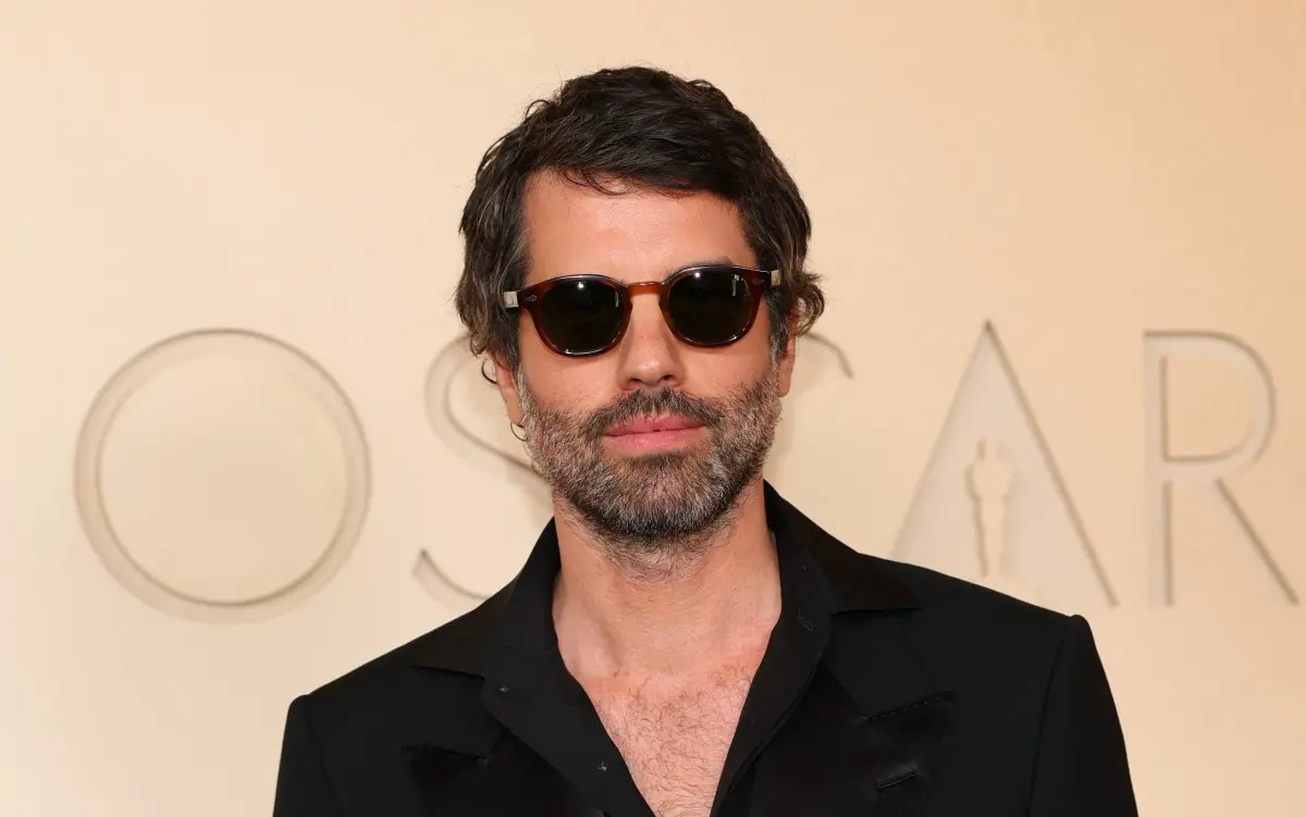 Pessoa posando em frente a um painel claro com a palavra “OSCAR” parcialmente visível ao fundo. A pessoa veste um traje preto com gola estruturada e abertura no peito. O cabelo é curto e levemente ondulado. A iluminação é suave e uniforme, destacando o contraste entre a roupa escura e o fundo claro.