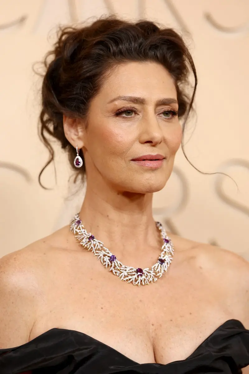 Foto de uma pessoa em close no tapete vermelho, usando um vestido preto de tecido estruturado que deixa os ombros à mostra. A pessoa usa um colar chamativo composto por peças entrelaçadas em tom metálico claro, com detalhes em roxo, combinando com um par de brincos pendentes que também apresentam pedra roxa. O cabelo está preso em um penteado com fios soltos, e o fundo exibe um painel claro associado ao evento do Oscar.