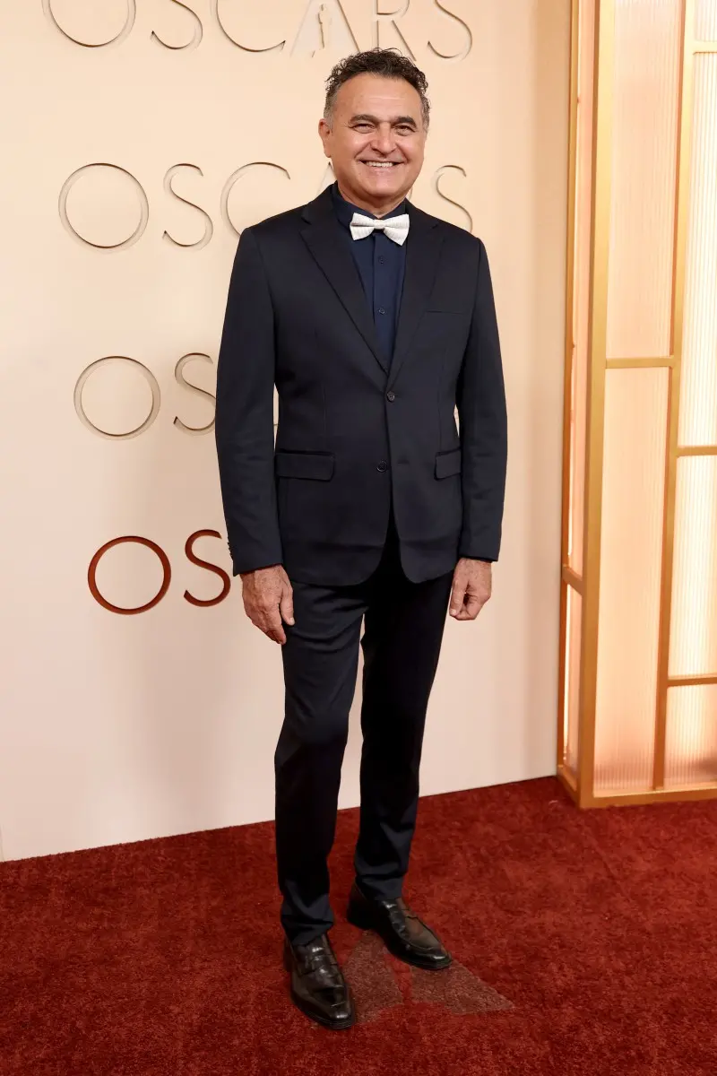 Pessoa em um tapete vermelho posando diante de um painel claro com a palavra “OSCARS” repetida ao fundo. A pessoa veste um terno escuro de corte clássico, composto por blazer, calça combinando e camisa azul-marinho.