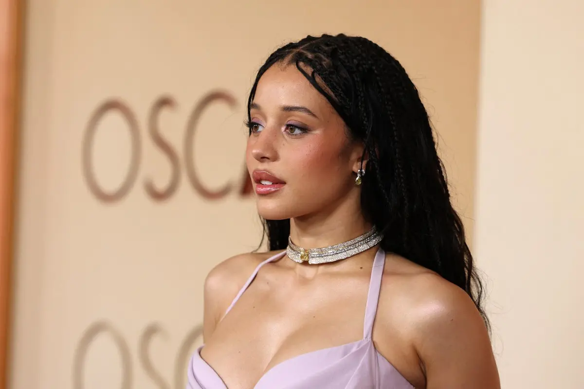 Pessoa em traje lilás com alças finas, usando colar brilhante e brincos, posando diante do painel do Oscar.