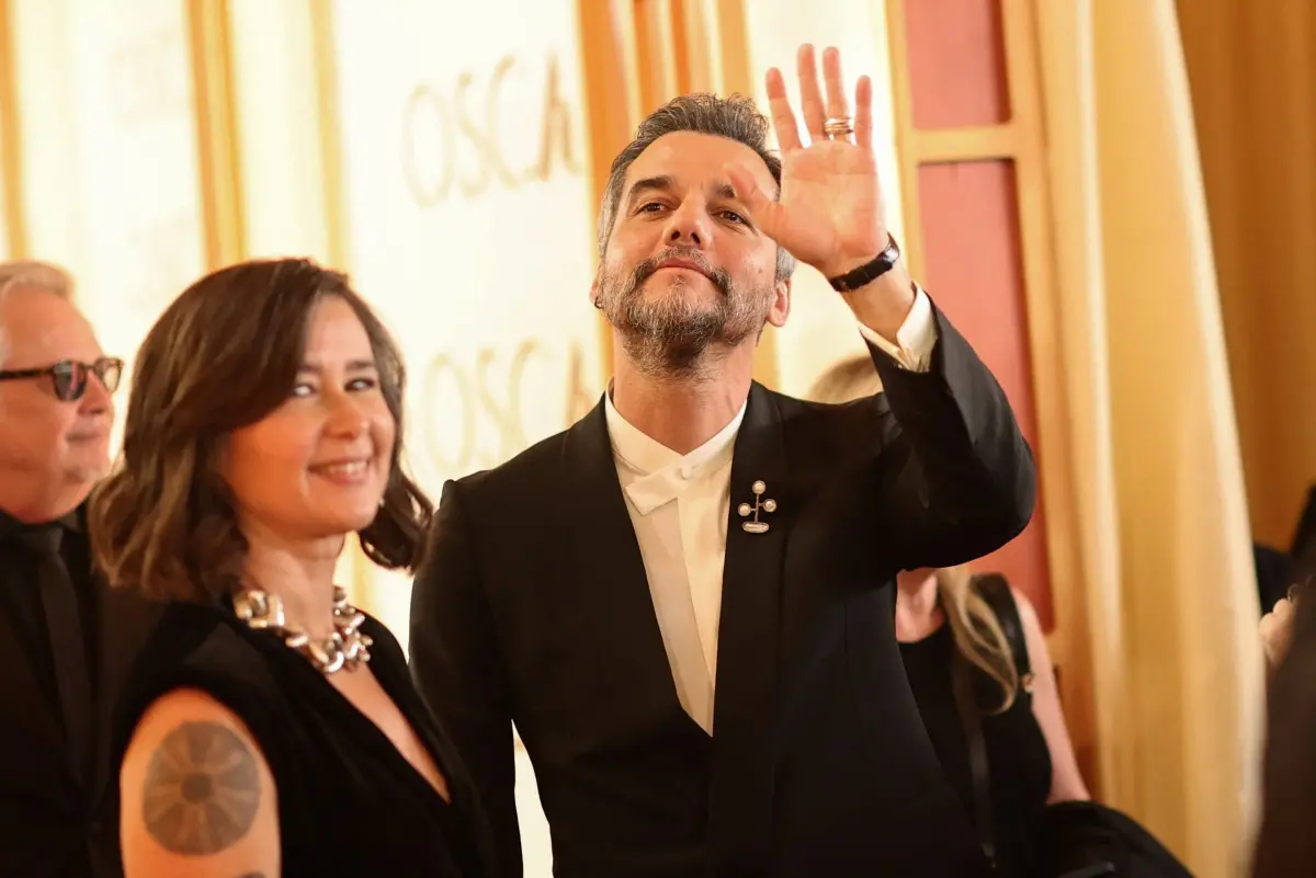 Duas pessoas aparecem em um ambiente de tapete vermelho iluminado por luz quente, com um fundo claro onde se lê “OSCARS” em grandes letras. A pessoa à direita veste um terno preto com camisa branca estruturada e um broche metálico abstrato preso ao paletó; essa pessoa ergue uma das mãos, exibindo anéis.