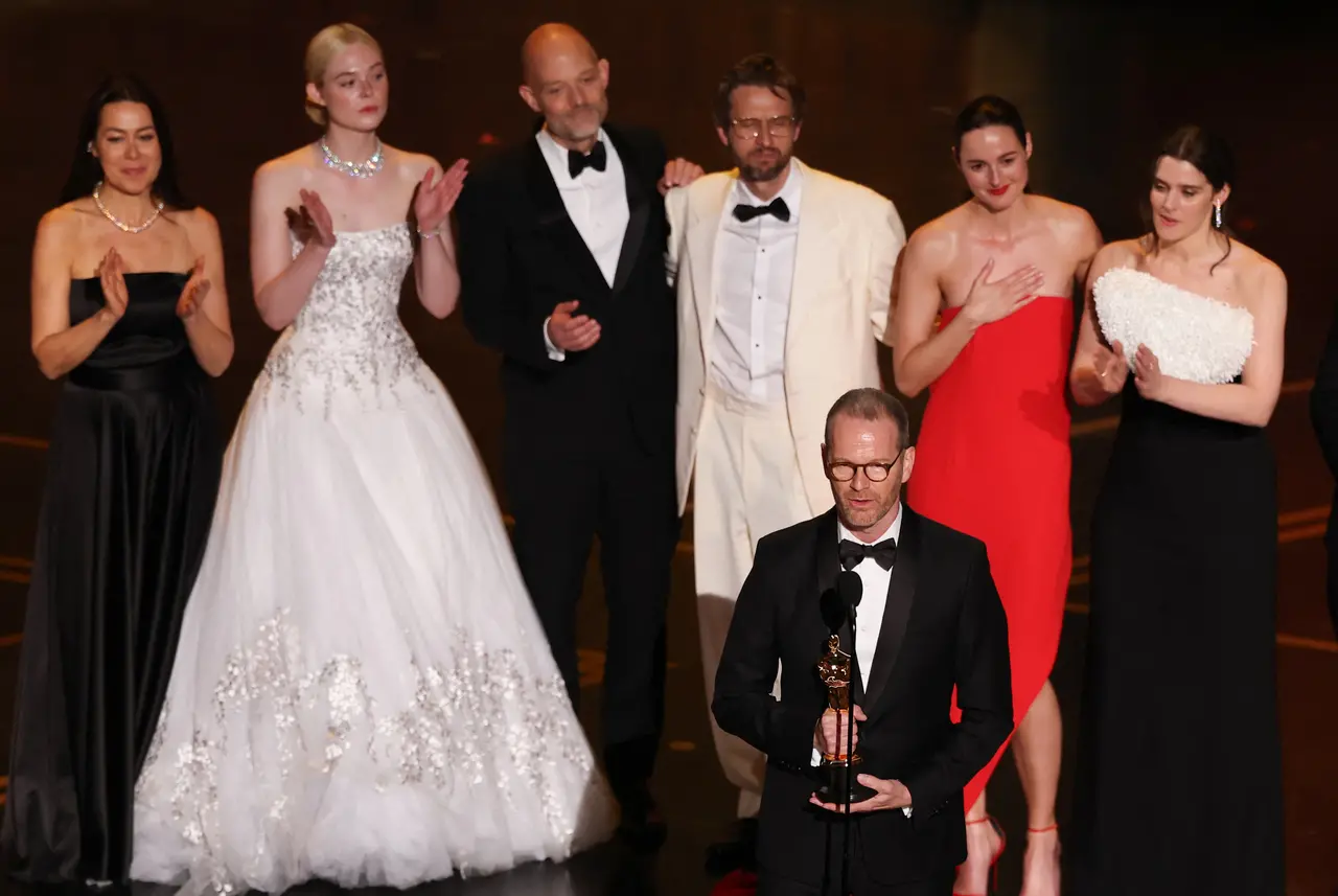 O cineasta Joachim Trier discursa no palco do Oscar 2026 segurando sua estatueta, enquanto o elenco de 