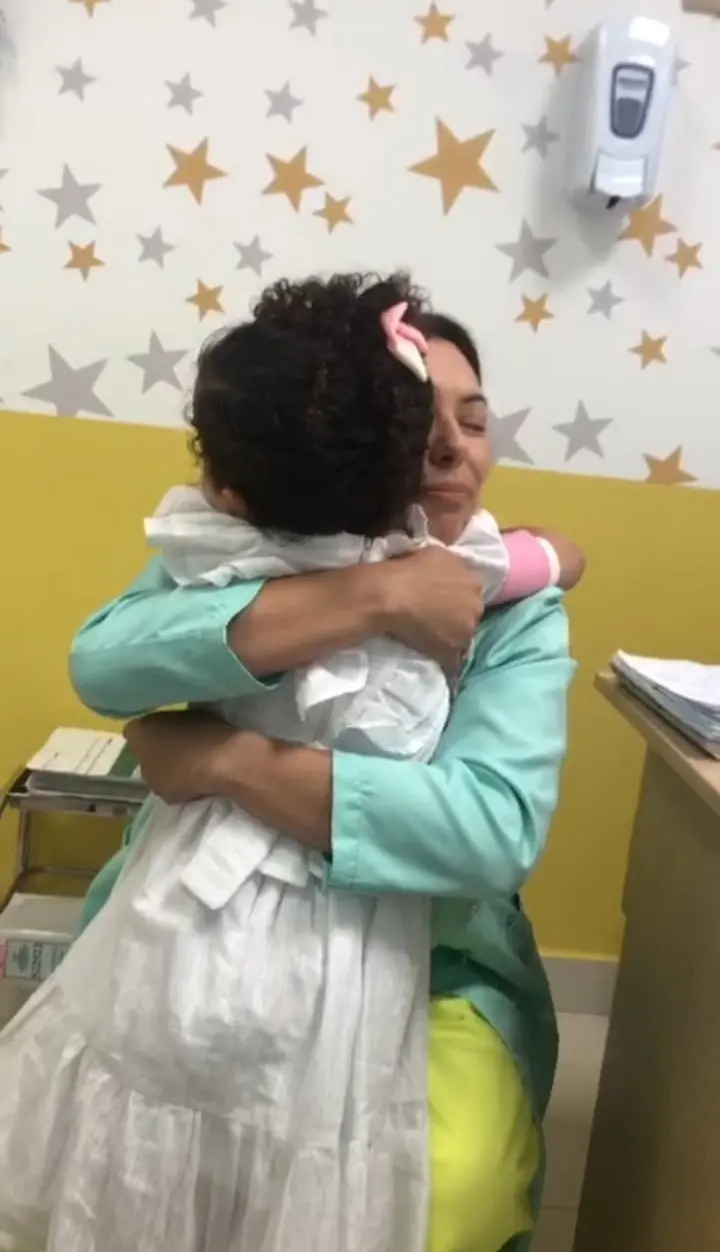Mulher em uniforme verde abraçando carinhosamente uma criança pequena de costas, em um ambiente com parede decorada com estrelas.