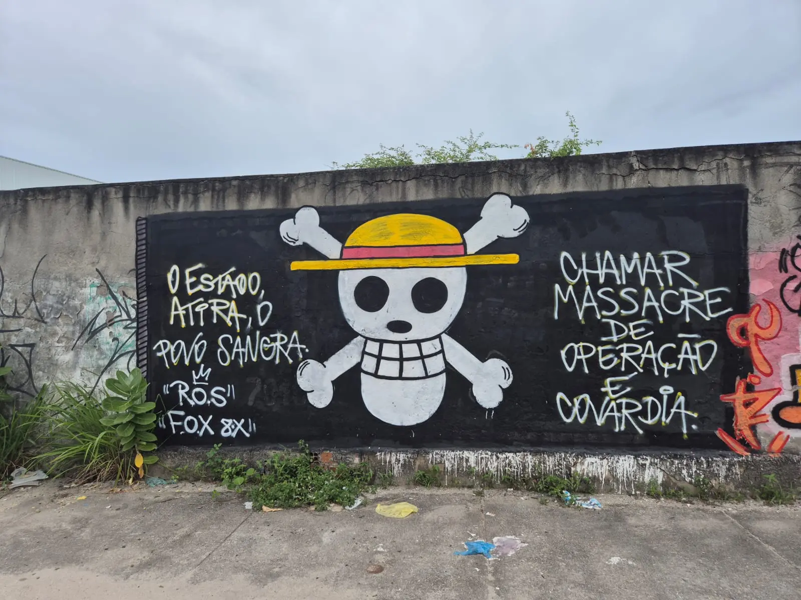 Grafite em muro preto com o símbolo pirata de One Piece (caveira com chapéu de palha) e duas mensagens de protesto em português: O Estado, atira, o povo sangra e Chamar massacre de operação é covardia.
