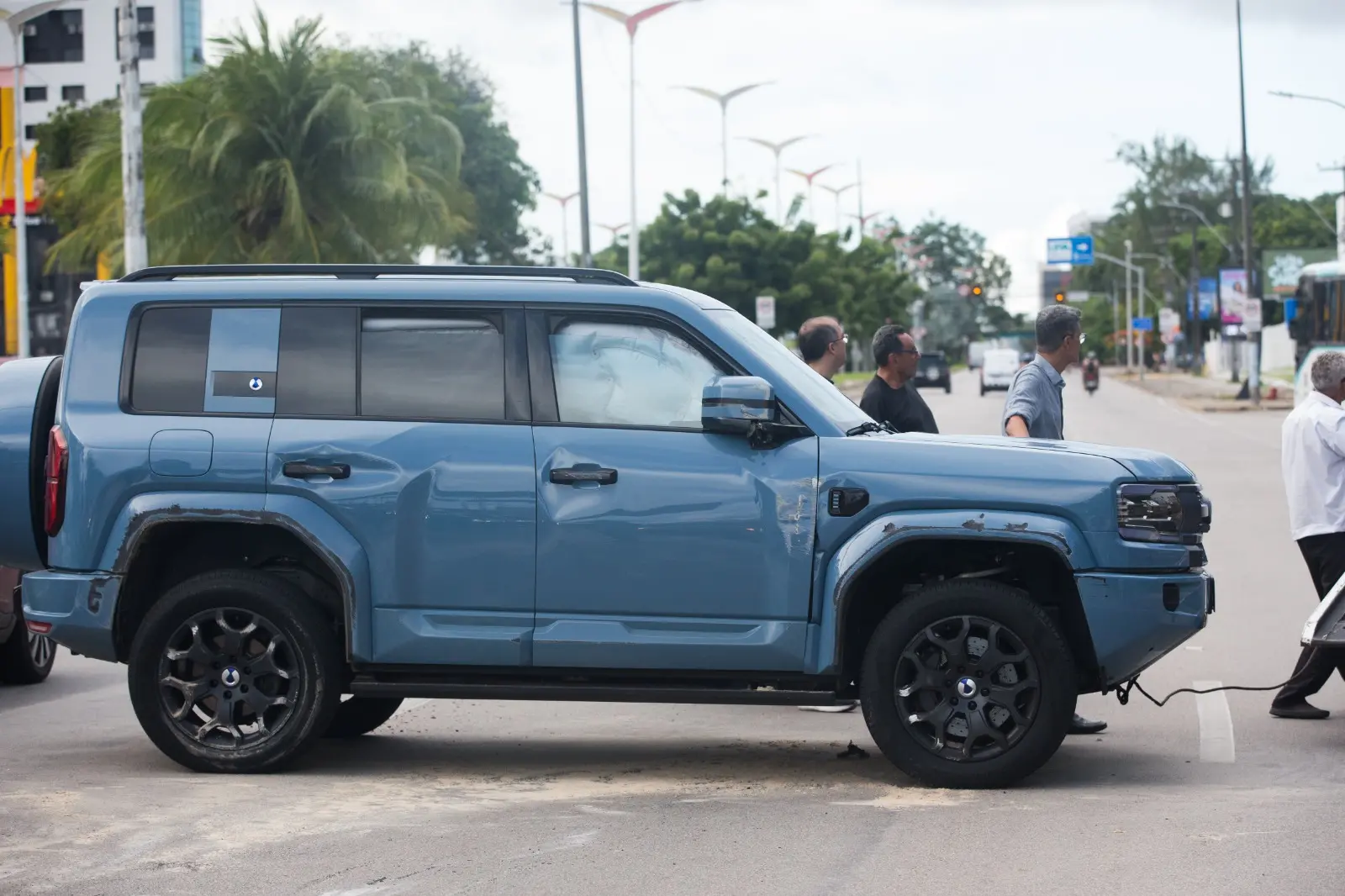 Um SUV azul claro apresenta danos na lateral e o airbag lateral acionado, enquanto homens observam o veículo em uma via pública. A composição foca no estado do carro após o impacto, com palmeiras e postes de iluminação ao fundo sob um céu nublado.