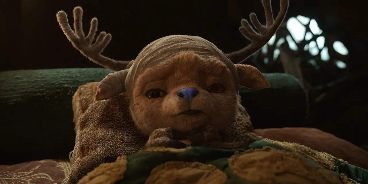 Uma pequena criatura de aparência peluda está deitada em uma cama com cobertores texturizados em tons terrosos. A criatura usa uma bandagem enrolada na cabeça e possui um par de chifres ramificados que se estendem para os lados. O ambiente ao redor é acolhedor e iluminado de forma suave, com elementos de madeira e um fundo levemente desfocado que sugere um interior rústico.