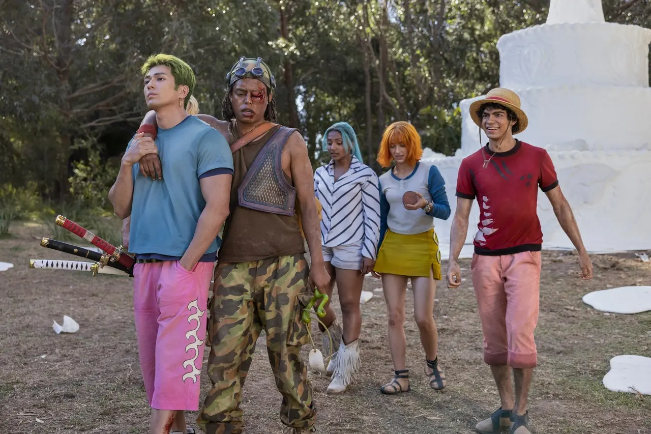 Um grupo de cinco pessoas caminha juntas em uma área ao ar livre cercada por árvores. Uma delas veste camisa azul e calças rosadas e carrega três espadas presas à cintura. Ao lado, outra pessoa usa roupas camufladas, uma camiseta marrom sem manga e um acessório na cabeça. Mais atrás, há uma pessoa com cabelo curto colorido, vestindo uma blusa listrada de manga comprida e shorts claros, e outra com cabelo laranja vibrante, usando uma blusa clara e uma saia amarela. À direita, uma pessoa com chapéu de palha e camisa vermelha caminha ao lado das demais. Ao fundo, aparece uma grande estrutura branca que se assemelha a um bolo cenográfico.