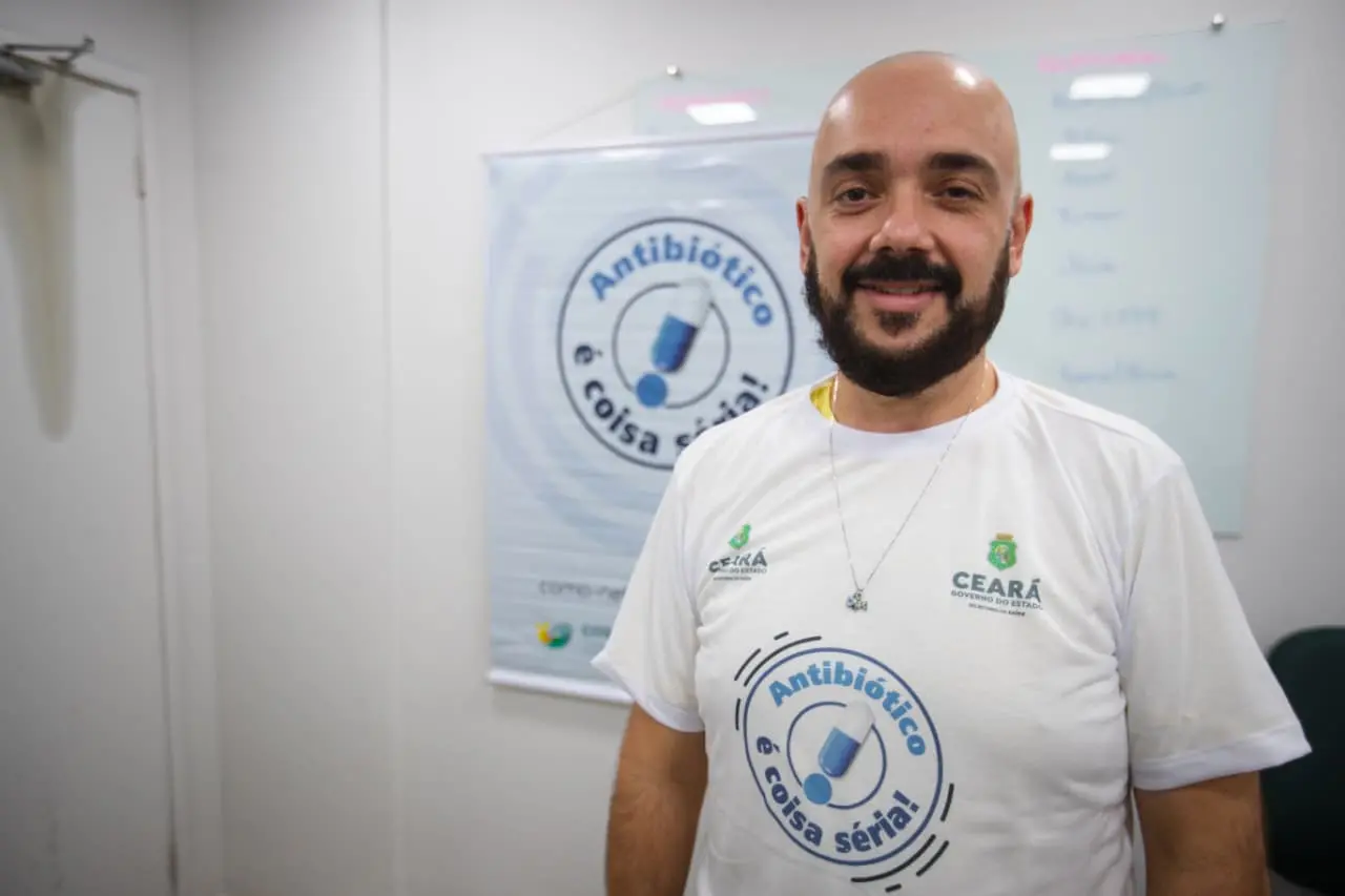 A imagem mostra o infectologista Lauro Perdigão Neto, secretário executivo de Atenção à Saúde e Desenvolvimento Regional da Sesa, vestindo camisa da campanha Antibiótico é Coisa Séria, com logo do Governo do Estado do Ceará e Secretaria da Saúde. Ele sorri em frente a banner da mesma campanha, ilustrando ações de conscientização sobre o uso racional de medicamentos.