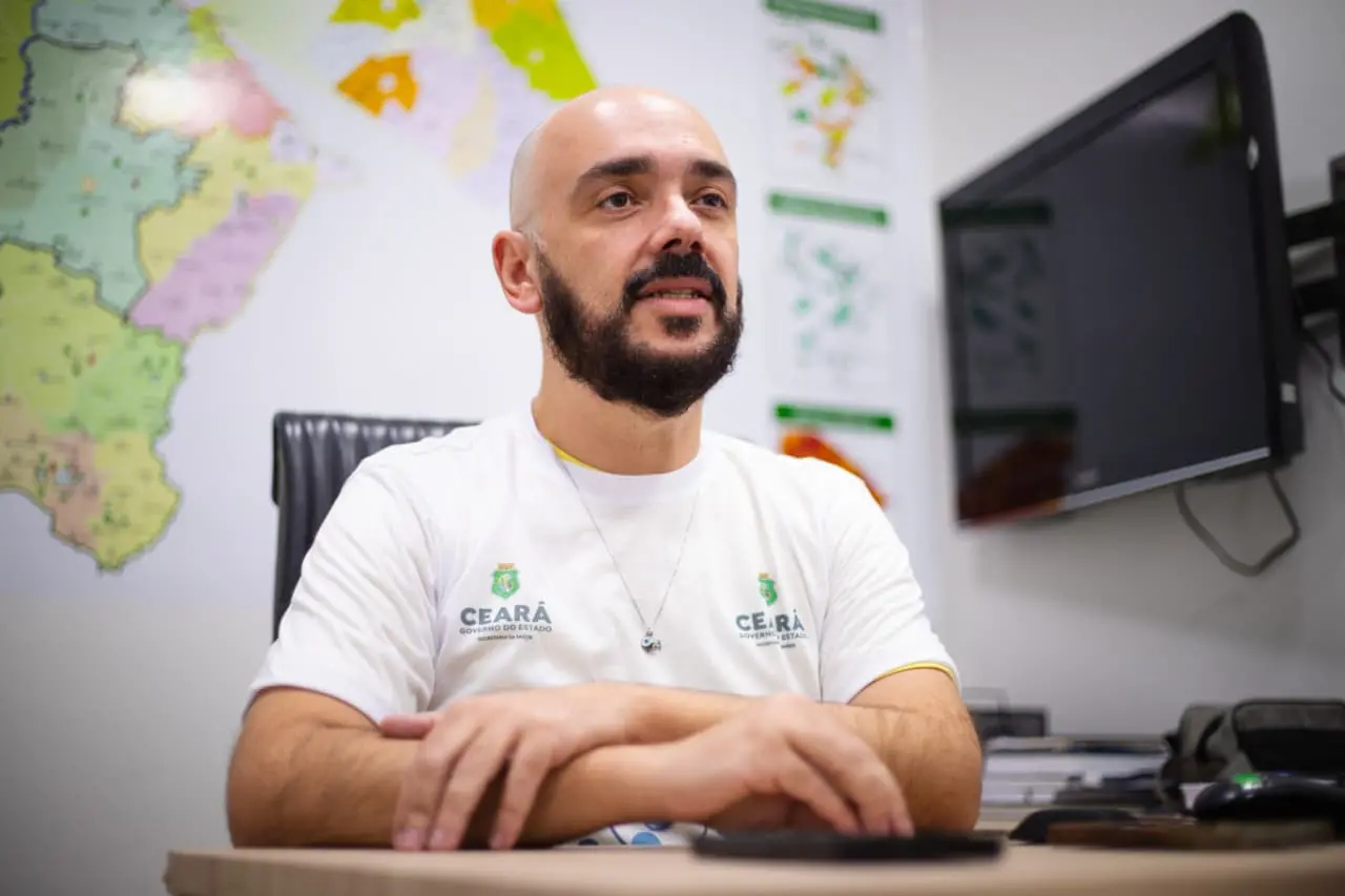 Imagem mostra o infectologista Lauro Perdigão Neto, secretário executivo de Atenção à Saúde e Desenvolvimento Regional da Sesa, durante entrevista em gabinete, vestindo camisa branca com o brasão do Governo do Estado do Ceará, tendo ao fundo mapas de monitoramento epidemiológico e indicadores de saúde.