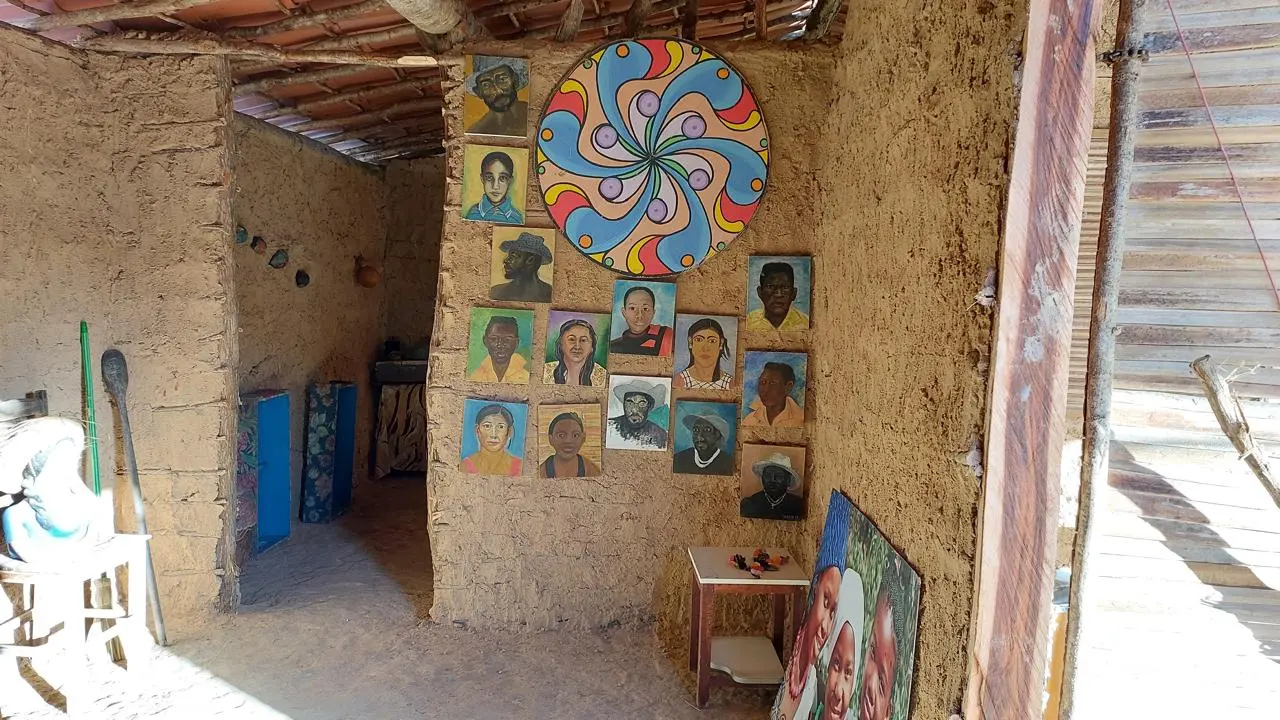 Galeria rural com paredes de barro exibindo pinturas de retratos coloridos. Uma peça abstrata redonda e sinuosa está pendurada acima. Ambiente rústico e luminoso.