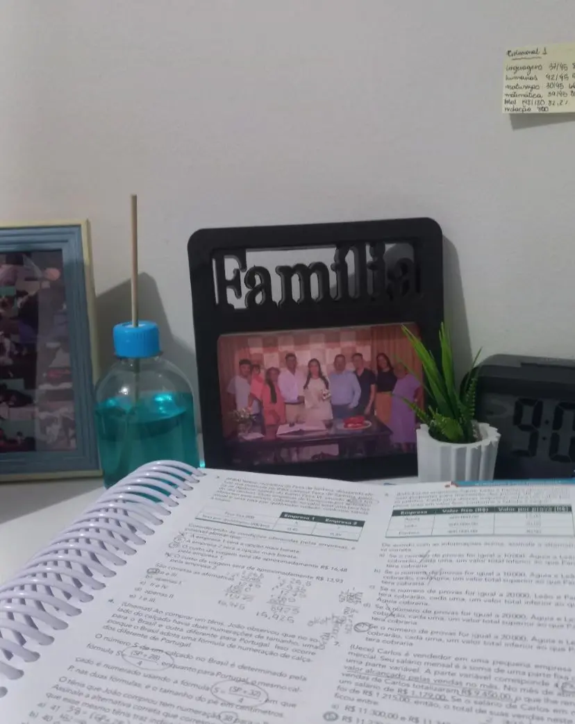 Uma apostila de estudos aberta em primeiro plano revela exercícios de matemática e anotações à mão em uma mesa de estudos. Ao fundo, um porta-retrato preto com a palavra 