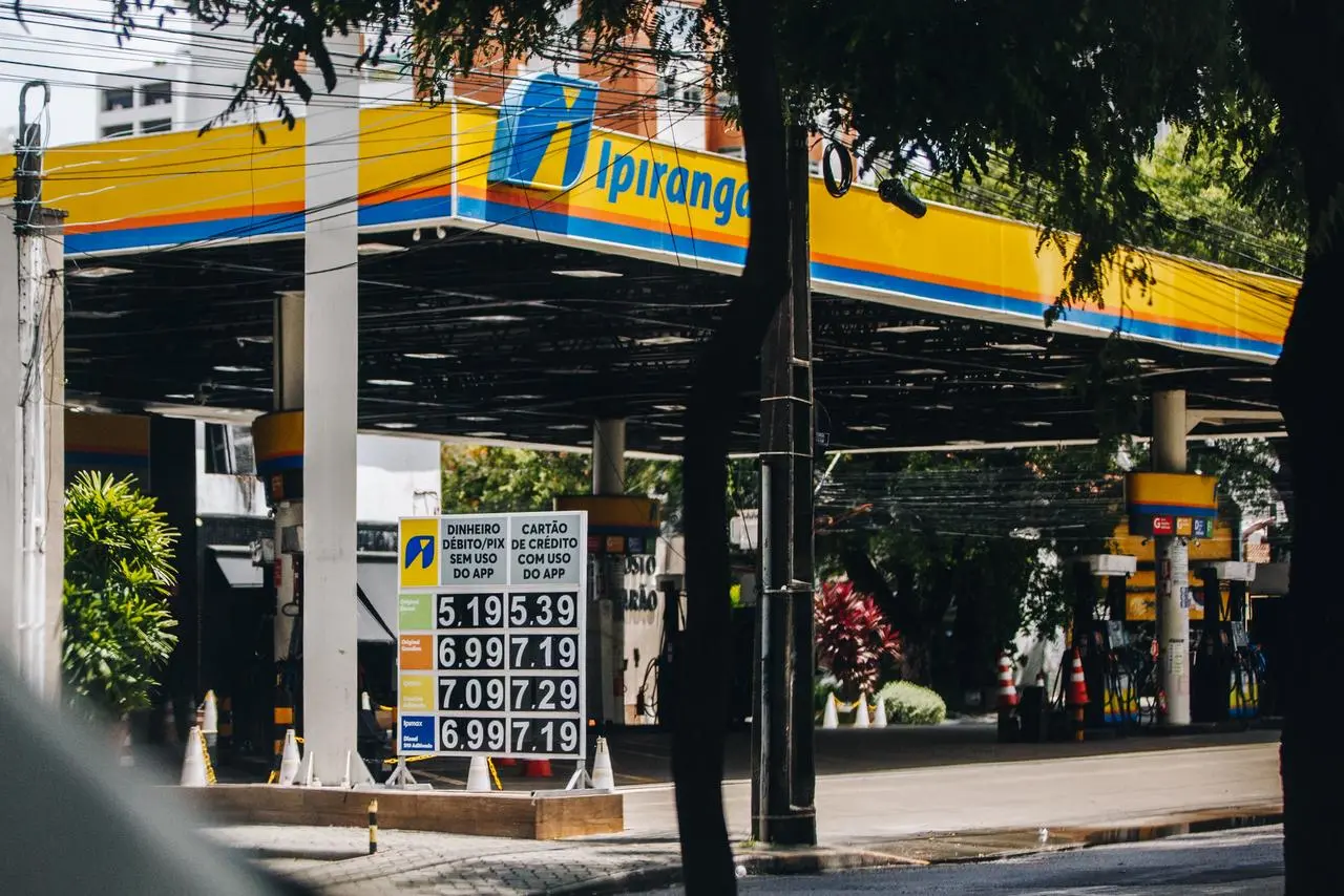Posto de gasolina da marca Ipiranga com placa exibindo preços dos combustíveis, visto da rua com árvores ao redor.