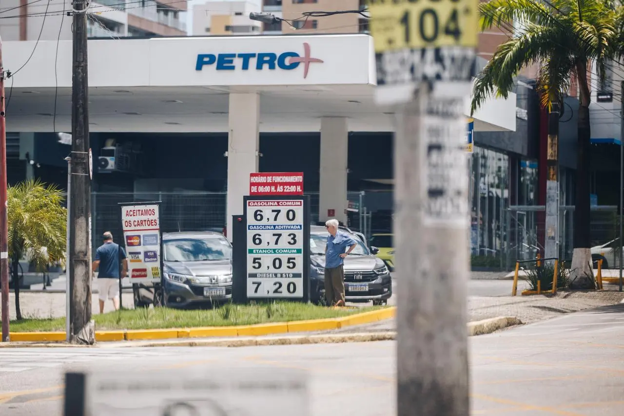 Posto de combustíveis da rede Petro+ com placa mostrando preços de gasolina, etanol e diesel, carros estacionados e pedestres ao redor.