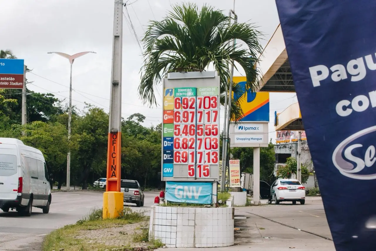 Placa de preços de combustíveis em posto às margens de avenida, com valores de gasolina, etanol, diesel e GNV, palmeira ao fundo e carros circulando.
