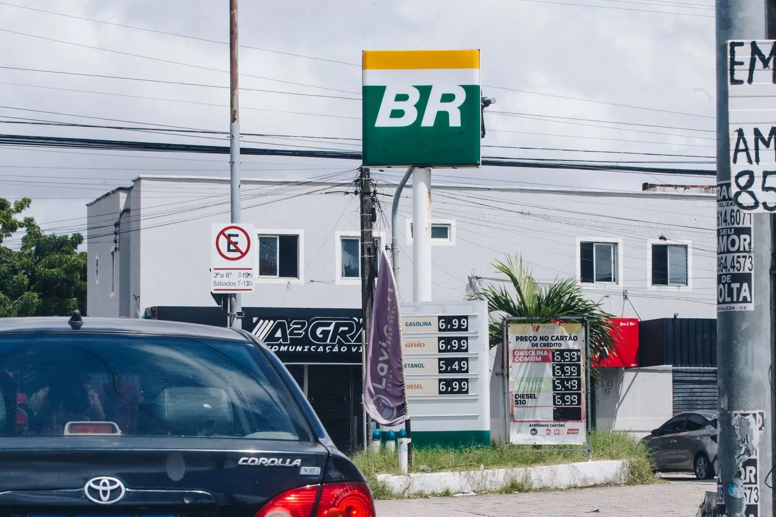 Placa de preços de combustíveis em posto com bandeira BR, exibindo valores de gasolina, etanol e diesel, com carros passando na via e prédios ao fundo.