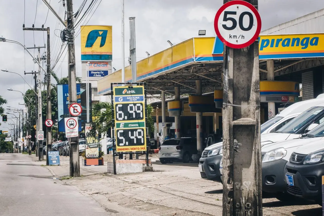 Posto de gasolina à beira da rua com placa de limite de 50 km/h e painel mostrando preços de combustível.