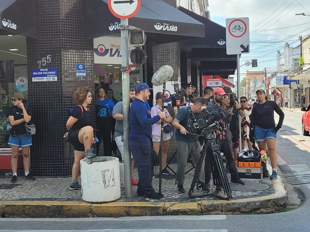 Câmeras, microfones e diversos profissionais ocupam uma esquina movimentada no Centro de Fortaleza durante as gravações de um longa-metragem durante o dia. A equipe se concentra ao redor do tripé da câmera em frente a uma loja, integrando a produção cinematográfica ao cotidiano urbano da cidade.