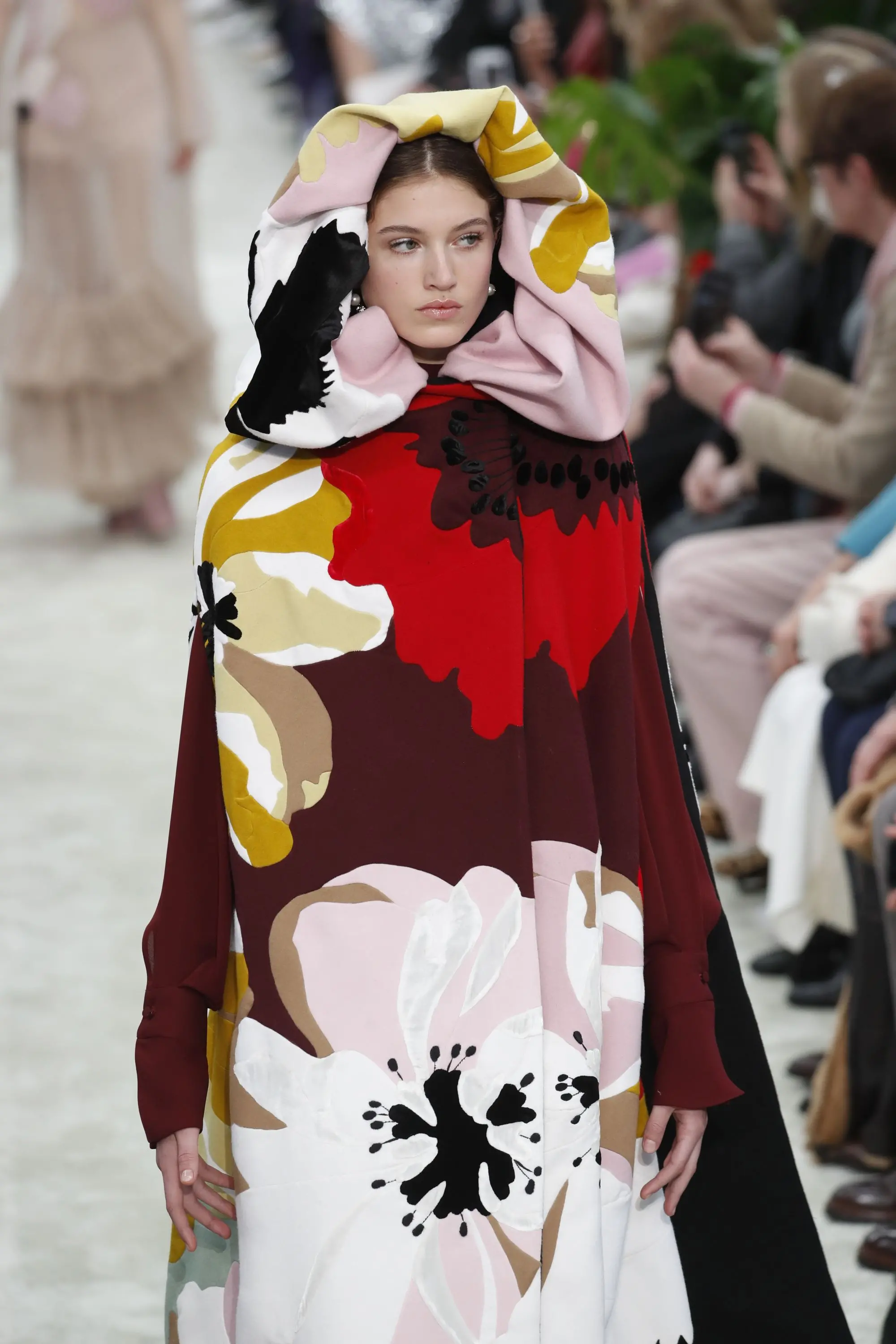 Modelo em desfile de moda vestindo uma capa ou casaco longo com capuz e estampas florais grandes e coloridas.