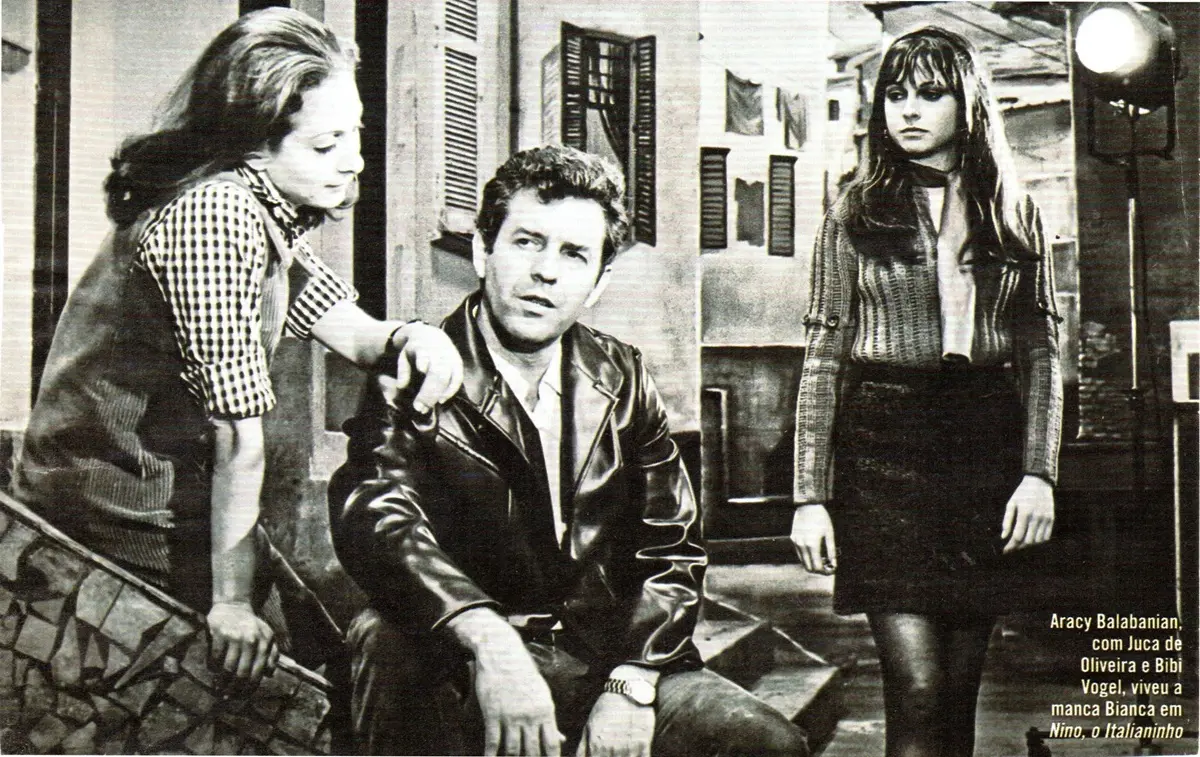 Cena em preto e branco de três atores em cenário urbano: um homem sentado ao centro, de jaqueta, conversa com uma mulher à esquerda, enquanto outra mulher, de pé à direita, observa a cena.