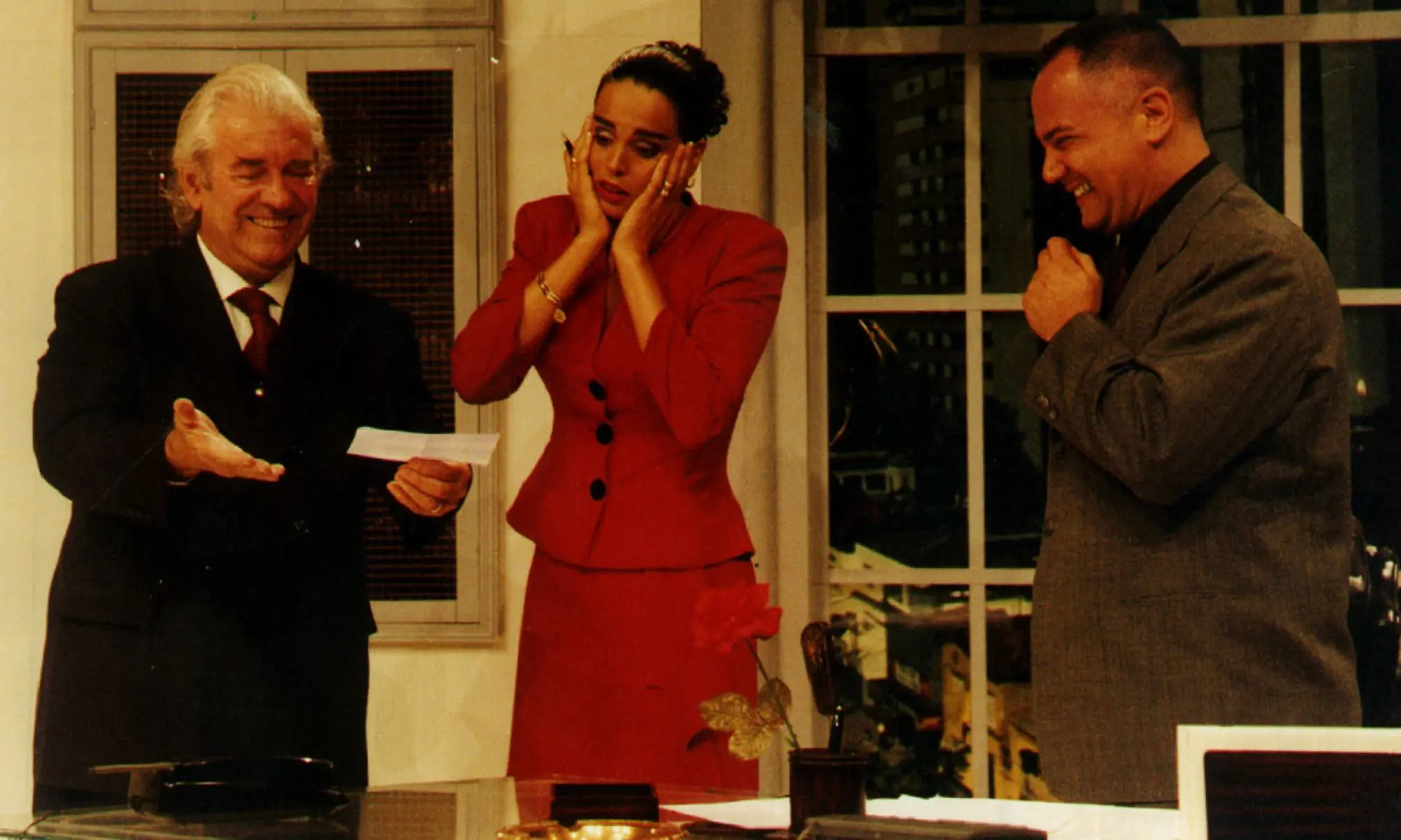 Cena teatral com três pessoas em um escritório: um homem mais velho de terno segura um papel e sorri, uma mulher de vermelho leva as mãos ao rosto em expressão de surpresa, e outro homem à direita ri, com a mão próxima ao rosto.