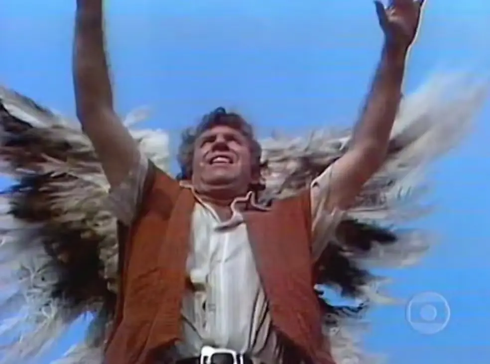 Homem com asas abertas ergue os braços para o céu, em expressão de êxtase, contra um fundo azul — cena de fantasia/novela.